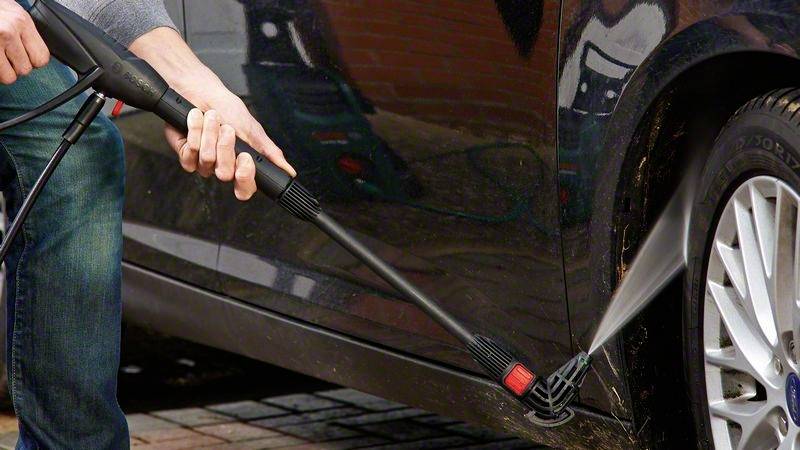 Person reinigt den unteren Teil eines Autos mit einem Hochdruckreiniger, sprüht Wasser auf den Reifenbereich, um Schmutz zu entfernen.