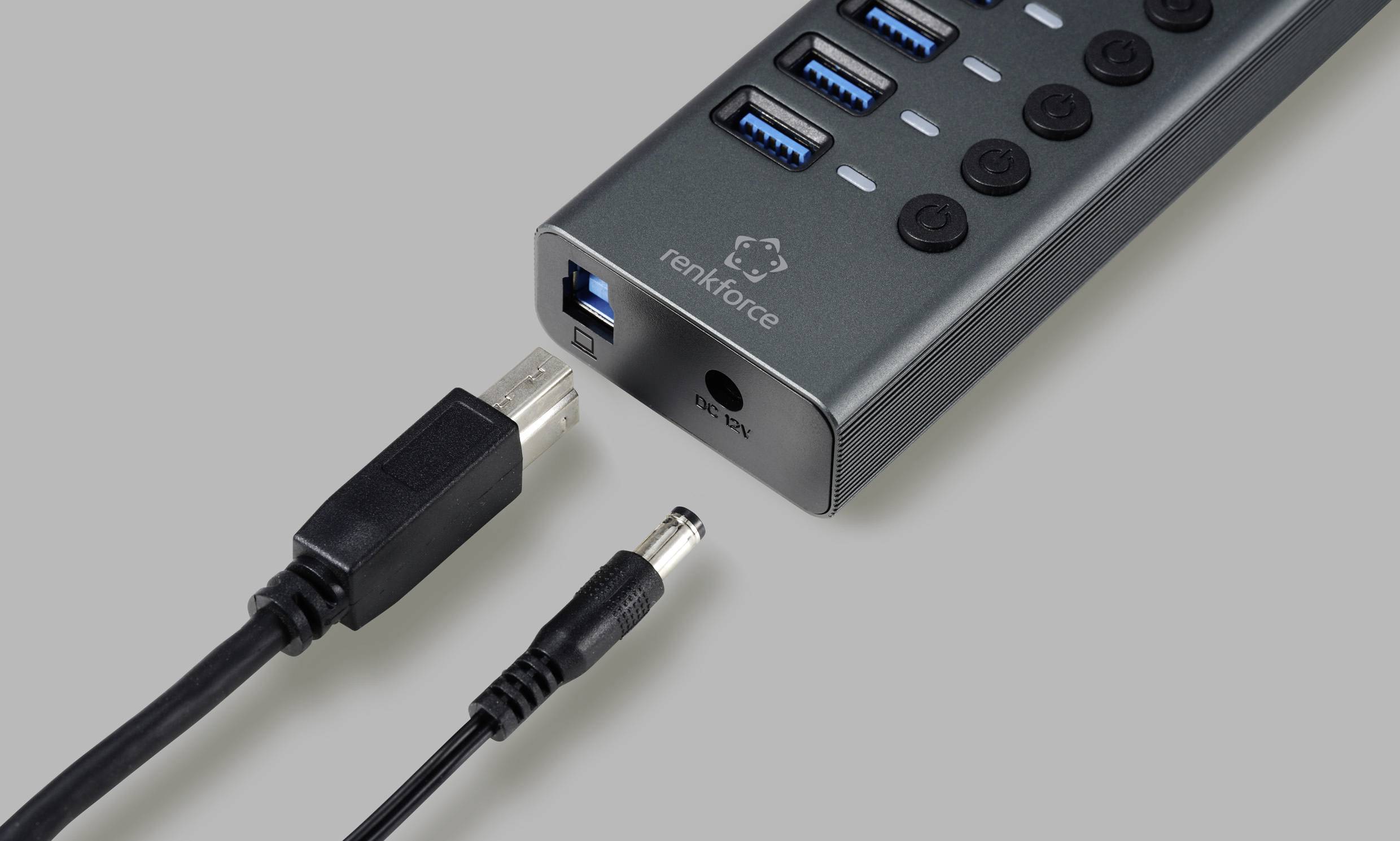USB-Hub mit vier USB-Anschlüssen und Ein-/Ausschaltern, Markenaufdruck 'renkforce', daneben zwei Kabel, USB und Strom, auf grauem Hintergrund.