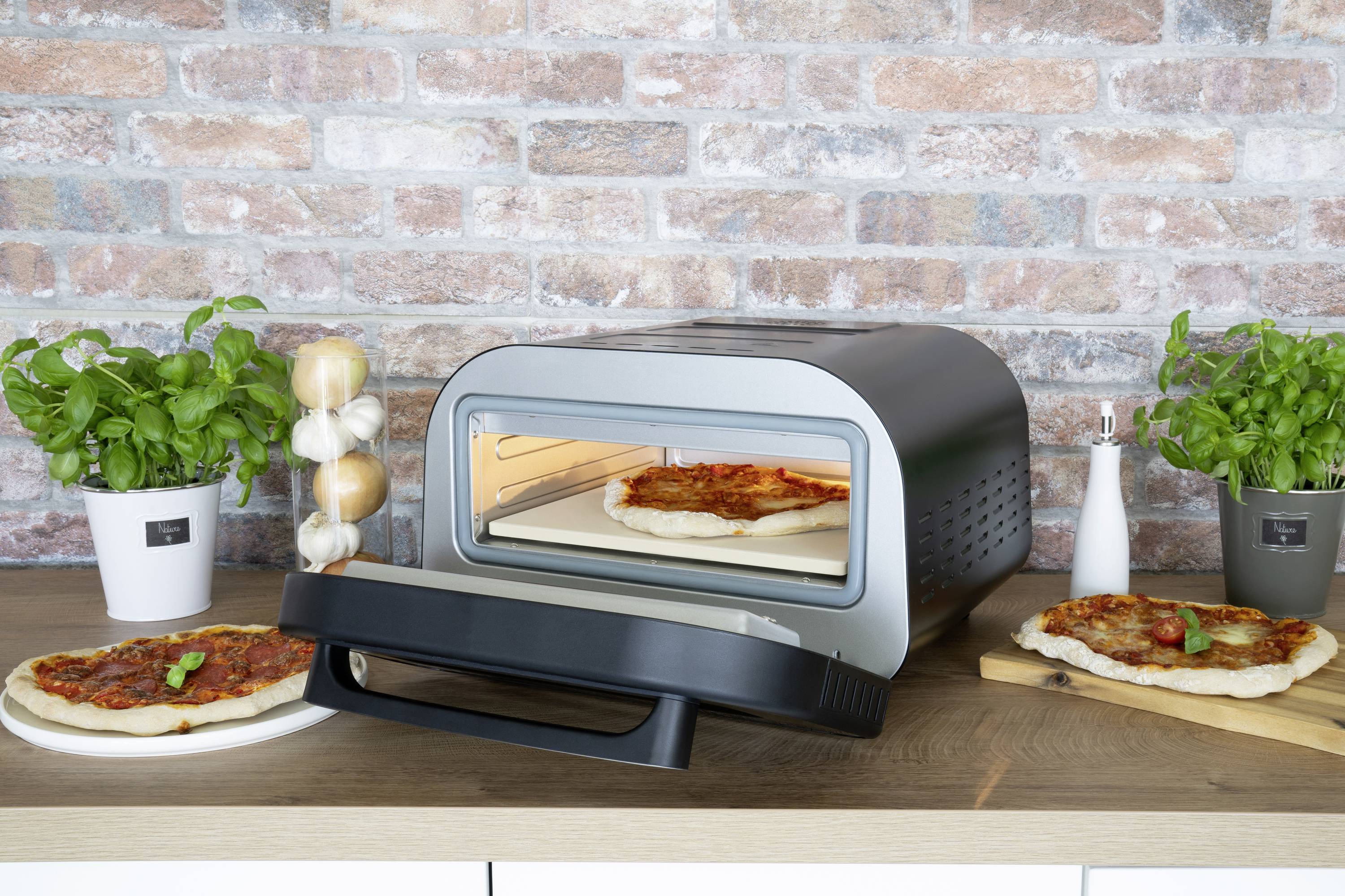 Ein elektro Pizzaofen mit geöffneter Tür, Pizza im Inneren. Zwei fertige Pizzen daneben, umgeben von frischen Kräutern und Gewürzen.