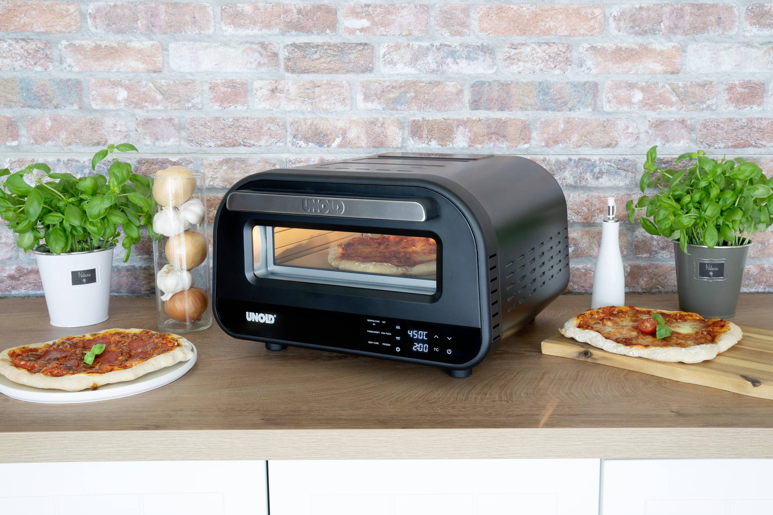 Ein schwarzer Pizzaofen steht auf einer Holztheke. Links und rechts daneben befinden sich zubereitete Pizzen, Kräutertöpfe und Gewürze.