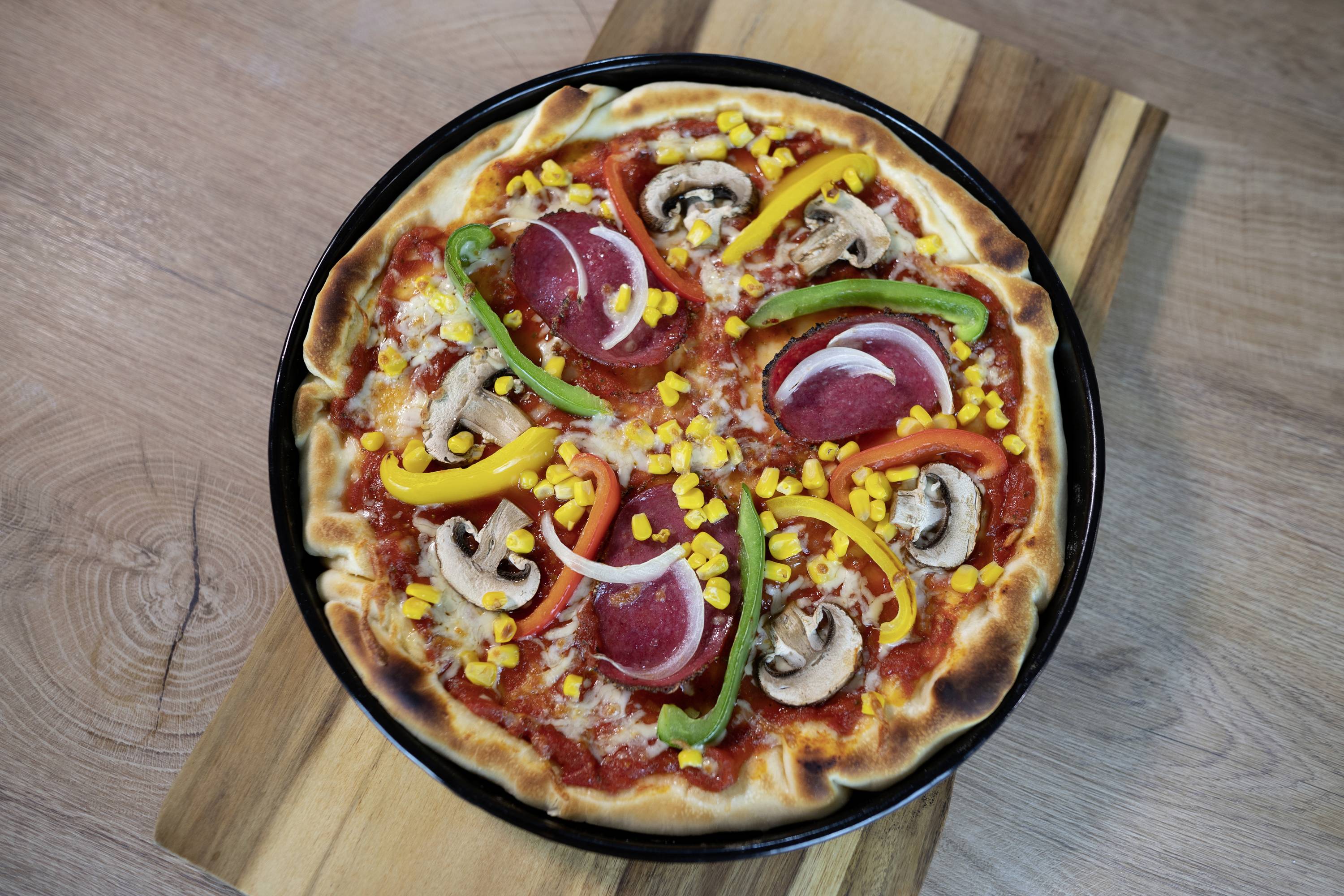 Pizza mit Tomatensoße, Peperoni, roten Zwiebeln, Paprika, Mais und Pilzen auf einem Holzbrett.