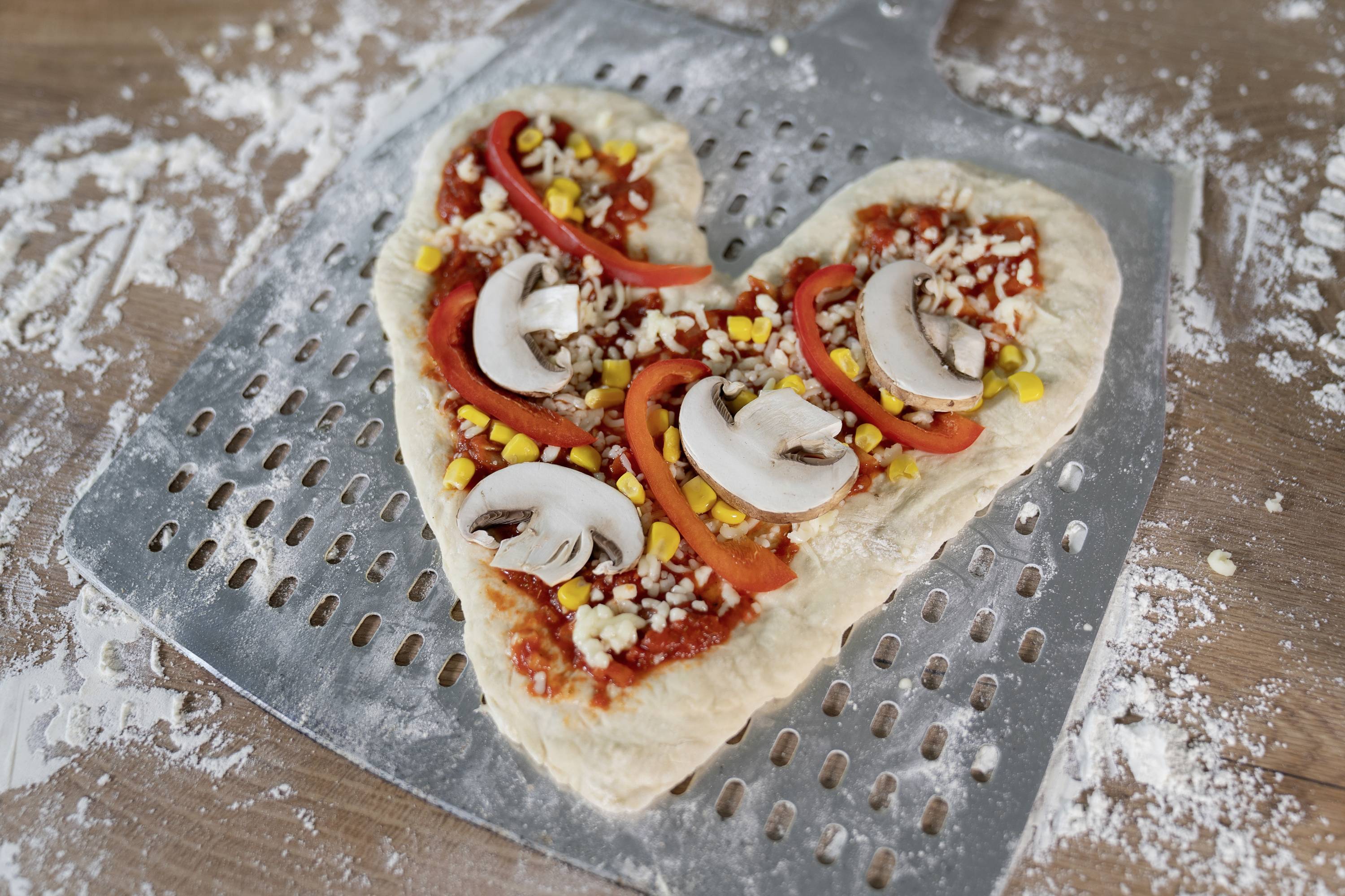 Herzförmiger Pizzateig mit Tomatensauce, Paprikastreifen, Champignons, Mais und Nüssen auf einer Pizzaschaufel.