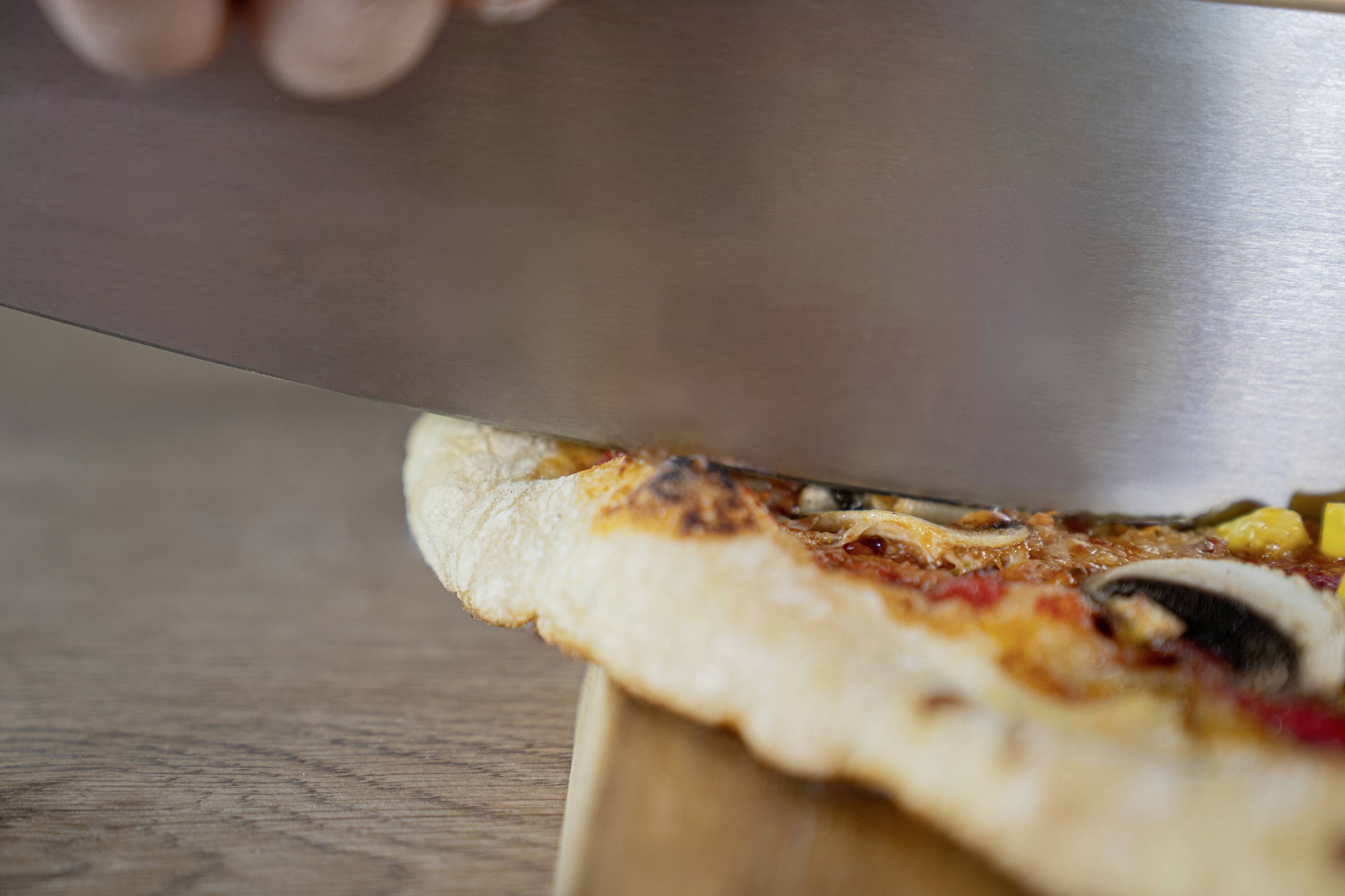 Nahaufnahme einer Pizza, die mit einem großen Messer geschnitten wird. Die Pizza hat Belag wie Pilze und Paprika.