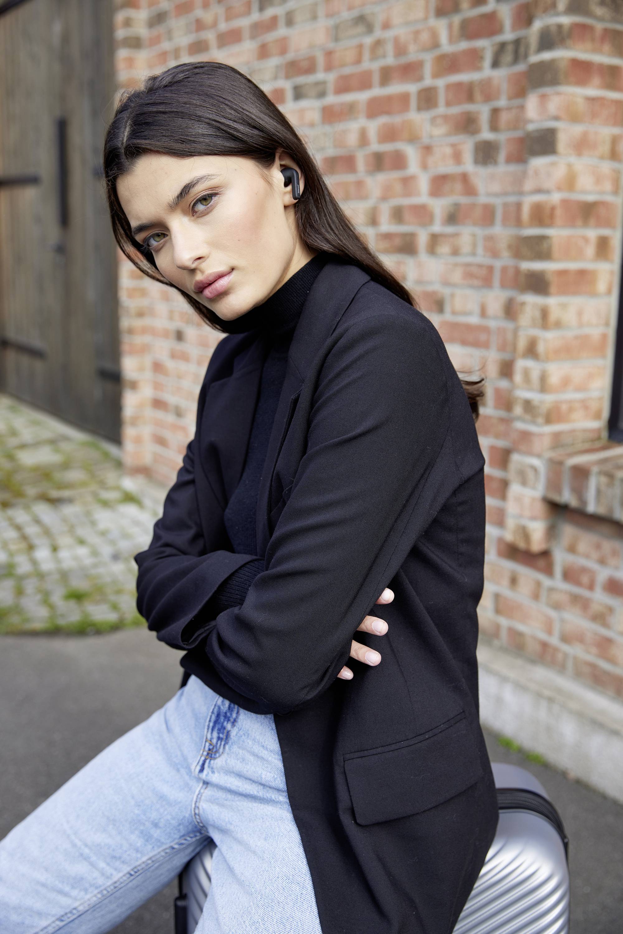 Frau in schwarzem Blazer und blauer Jeans lehnt lässig an einer Ziegelwand. Sie trägt drahtlose Ohrstöpsel und schaut ernst.