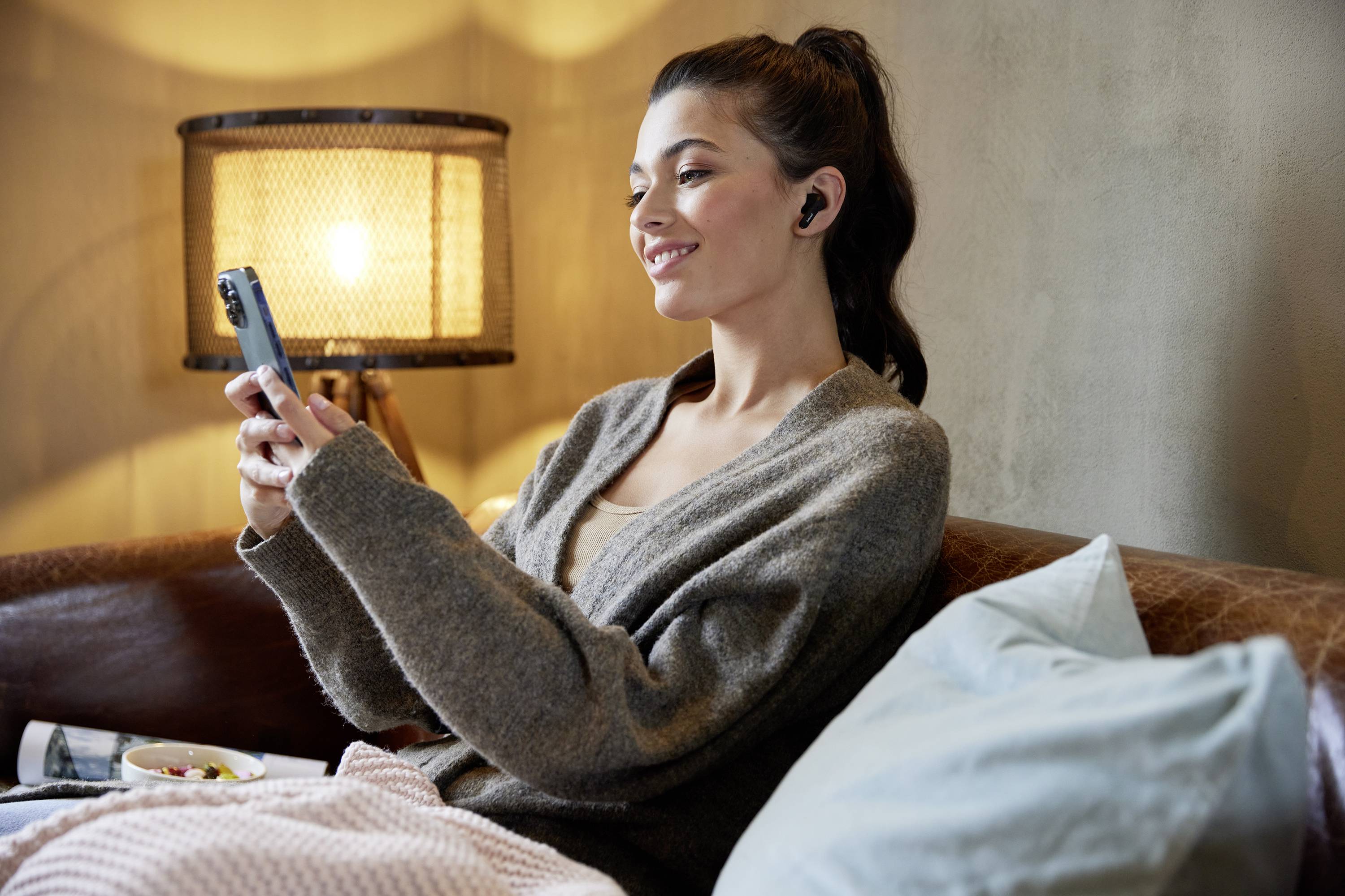 Eine lächelnde Frau sitzt auf einem Sofa, trägt kabellose Ohrhörer und schaut auf ihr Smartphone. Eine Lampe leuchtet im Hintergrund.