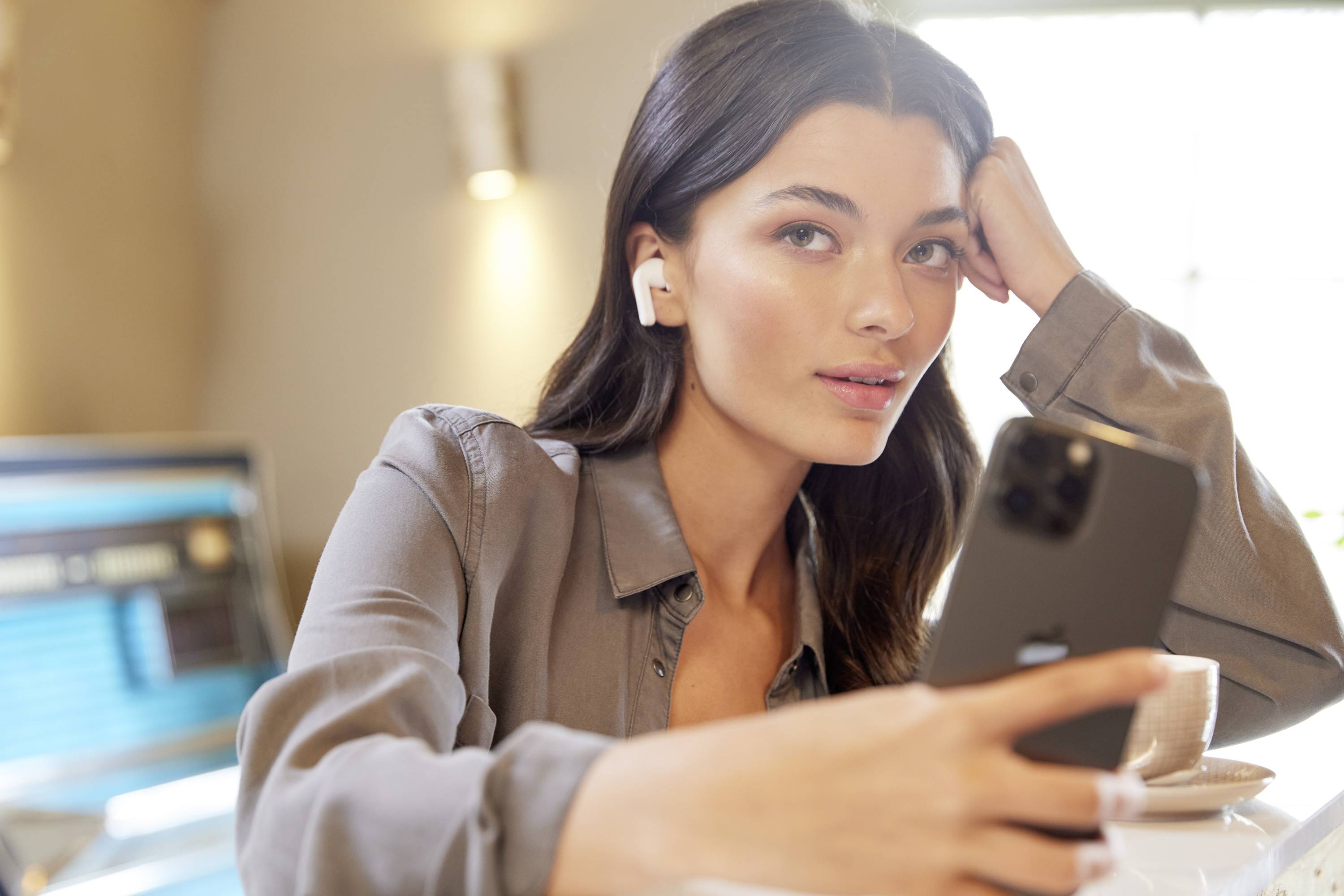 Eine Person sitzt an einem Tisch, hält ein Smartphone und trägt kabellose Ohrhörer. Im Hintergrund ist ein Computerbildschirm sichtbar.