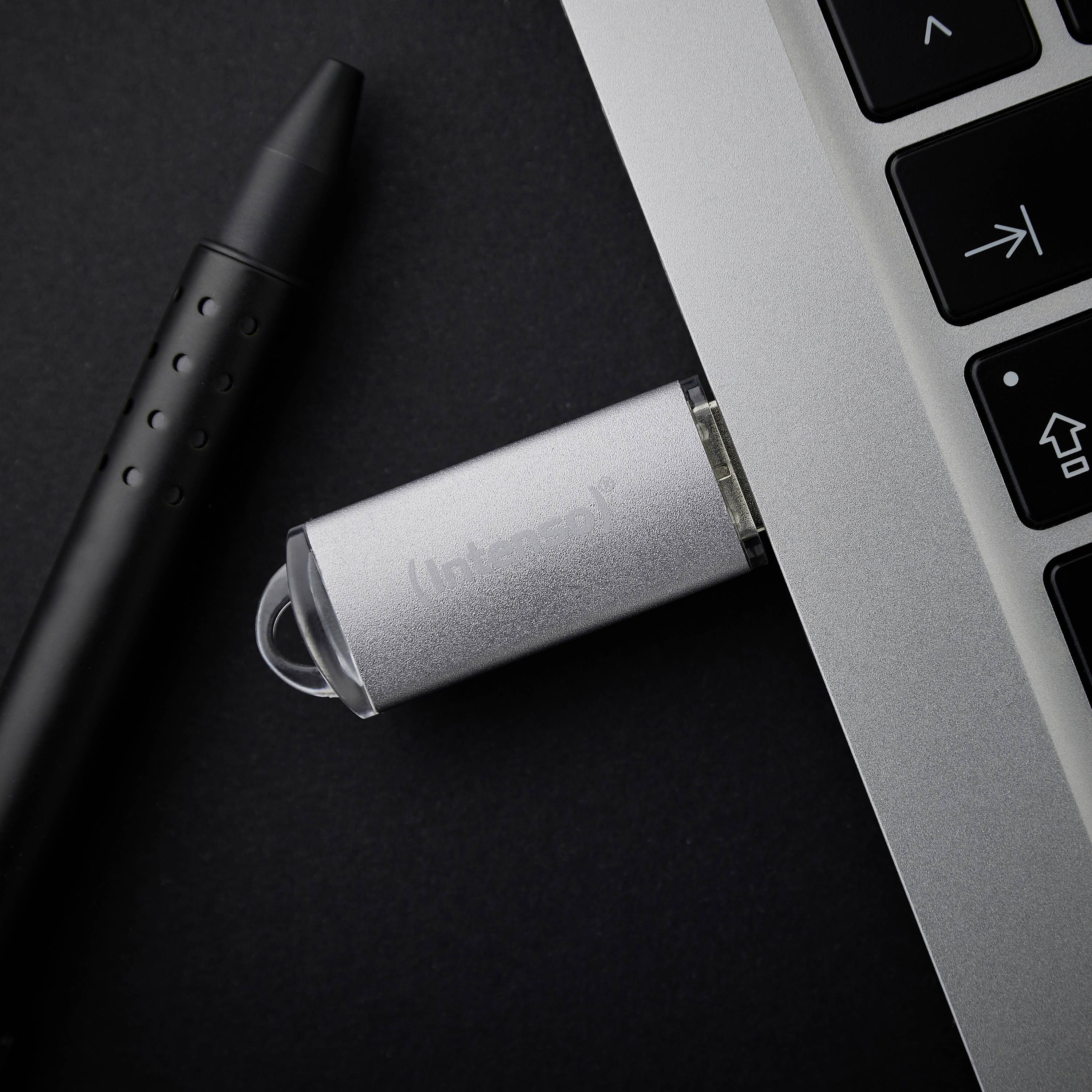 Ein silberner USB-Stick ist an einen Laptop angeschlossen. Daneben liegt ein schwarzer Stift mit Touch-Spitze auf einem dunklen Hintergrund.