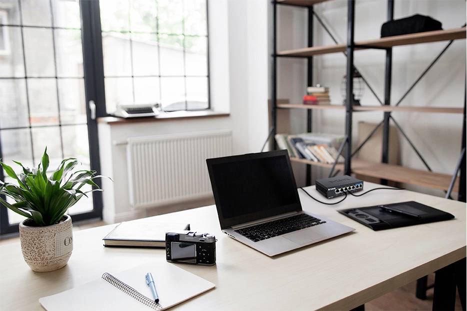 Ein aufgeräumter Schreibtisch mit einem Laptop, Notizbuch, Stift, Kamera und Pflanze. Im Hintergrund stehen Bücherregale.