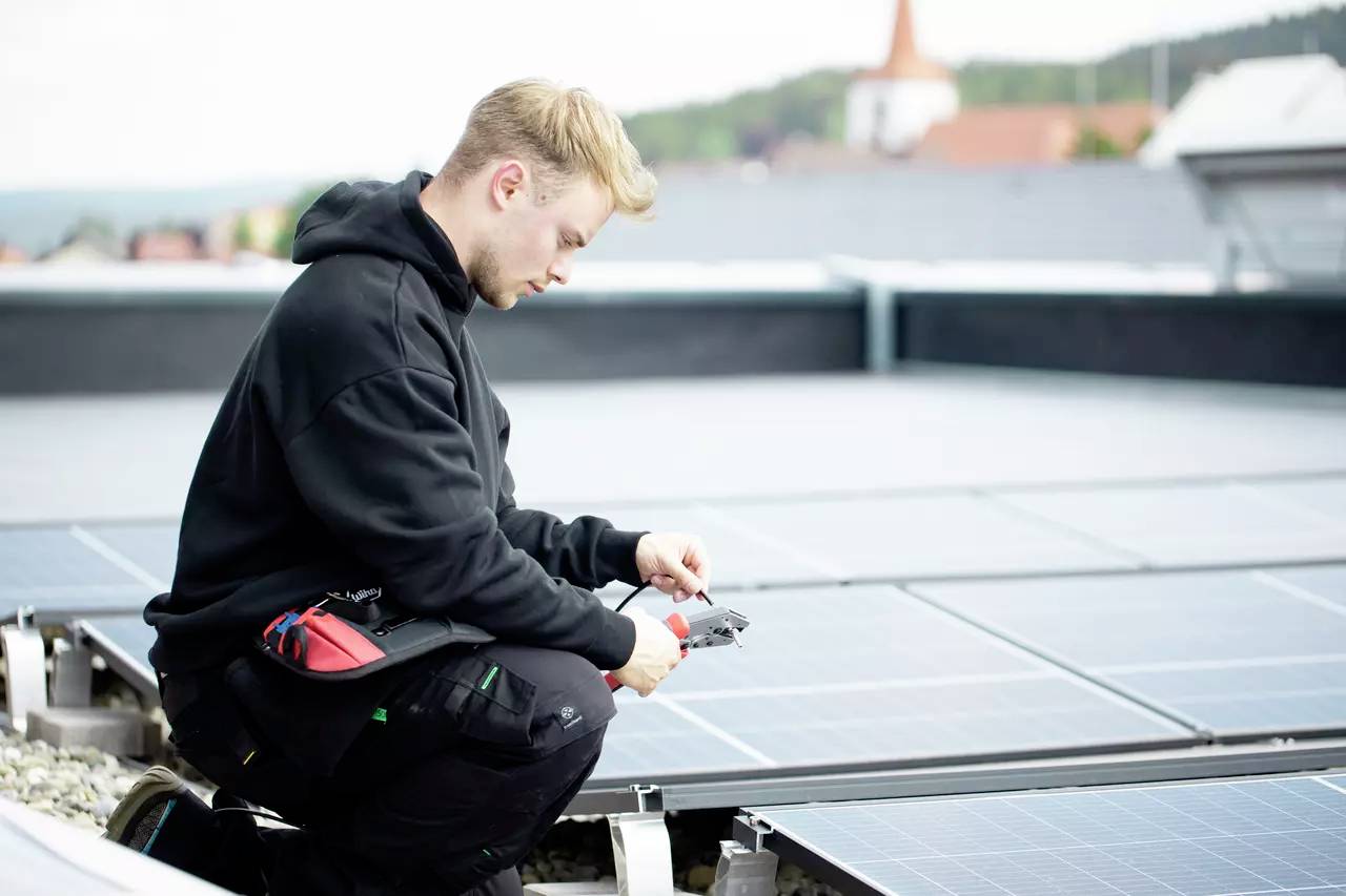 Eine Person in Arbeitskleidung prüft auf einem Dachkonstrukt Solarzellen mit einem Messgerät. Im Hintergrund sind verschwommene Gebäude zu sehen.