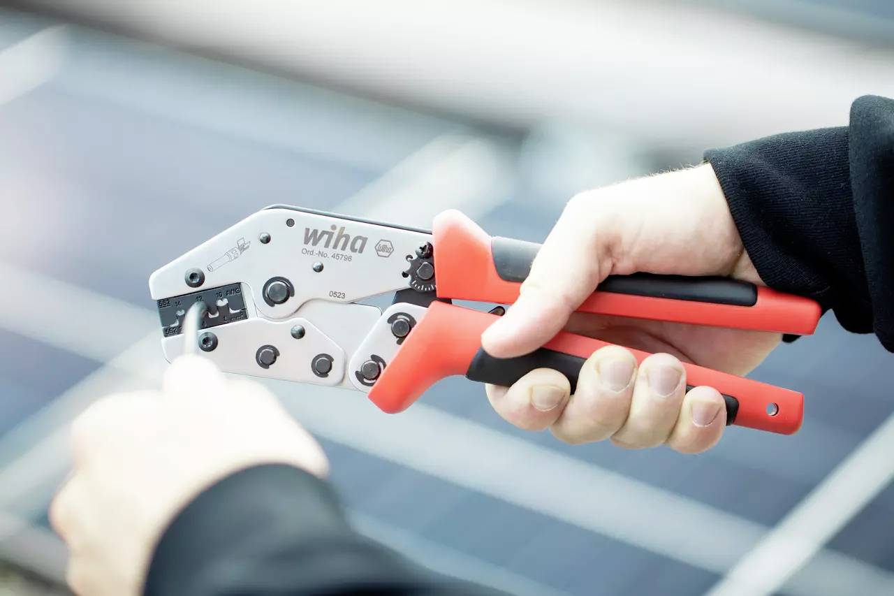 Eine Person hält eine rote Crimpzange über einem Solarpanel. Die Zange wird für elektrische Arbeiten verwendet.