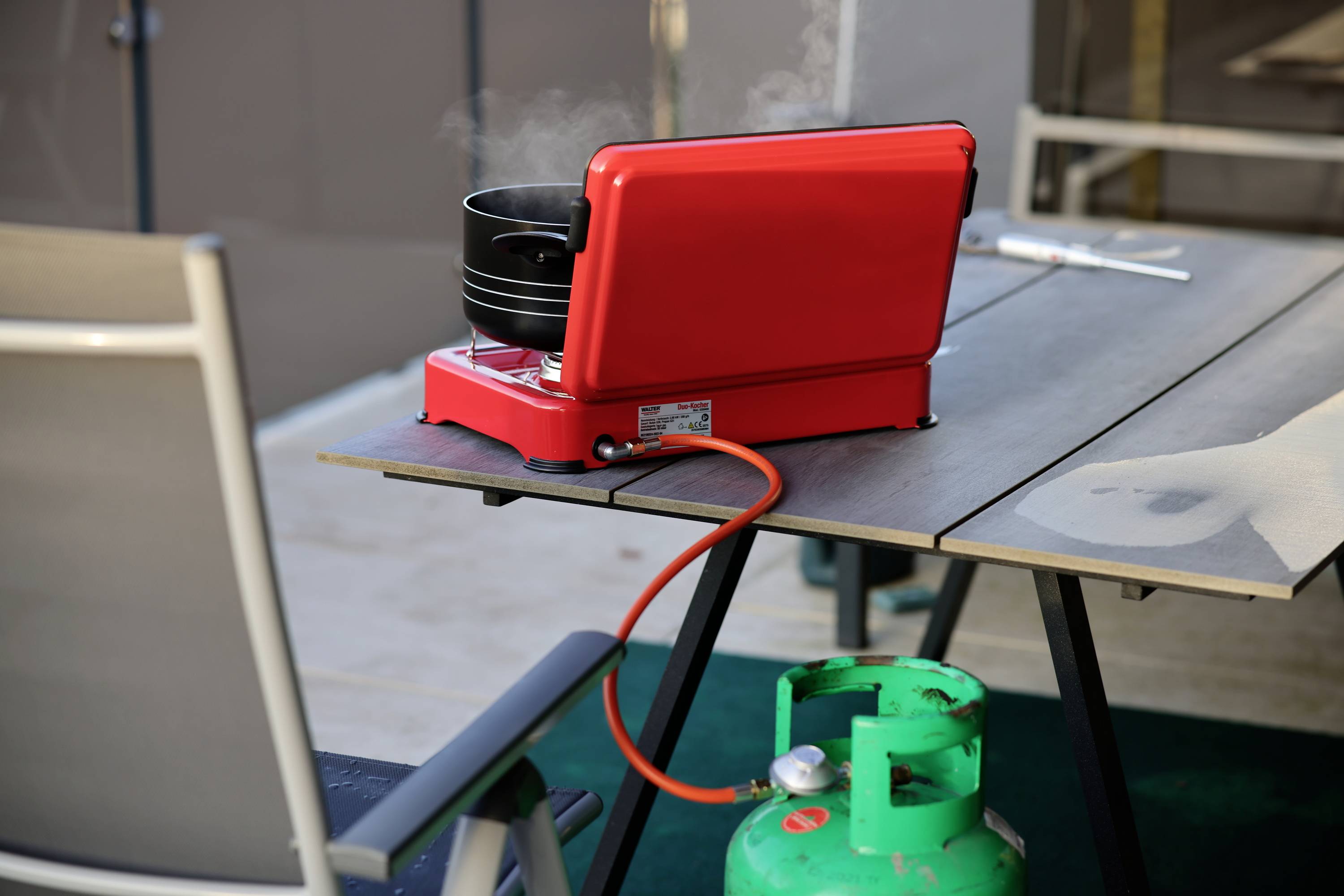 Ein roter Campingkocher mit Topf ist auf einem Tisch im Freien platziert. Eine Gasflasche darunter versorgt ihn mit Brennstoff.