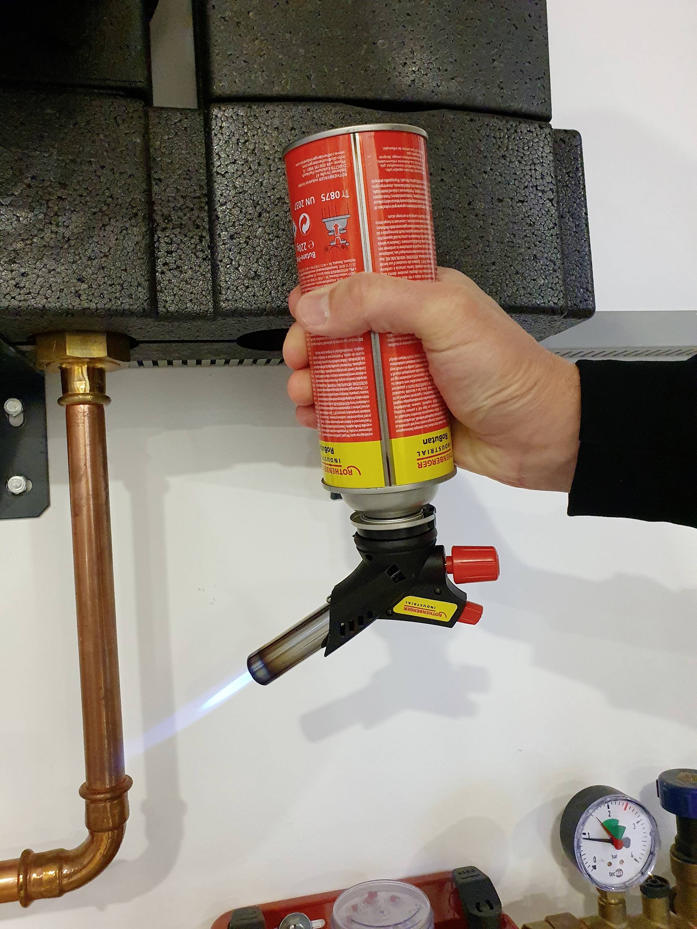 Eine Hand hält einen Gasbrenner mit blauer Flamme an einem Kupferrohr. Neben dem Rohr befindet sich ein Manometer.
