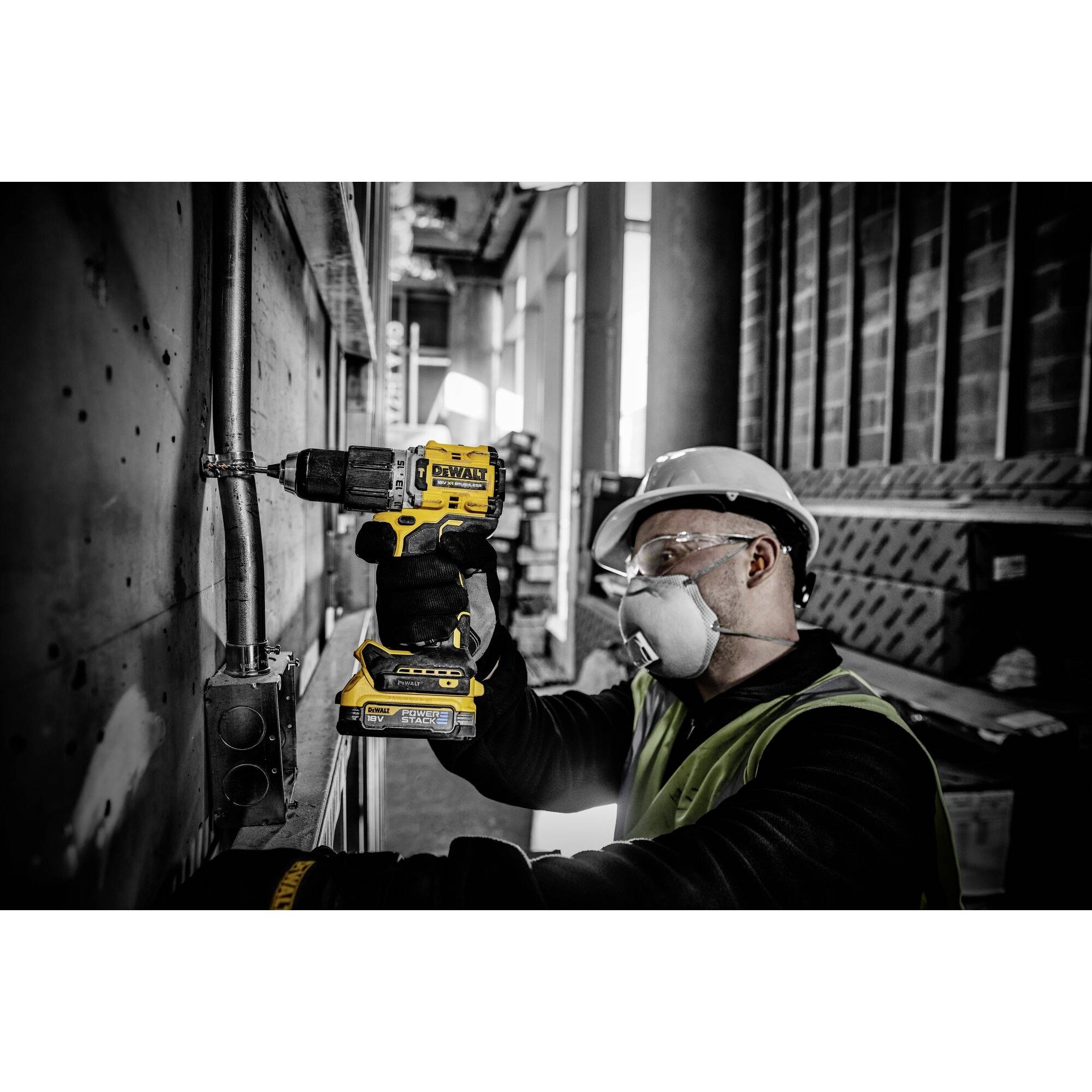 Arbeiter mit Schutzhelm und Maske verwendet eine gelbe Bohrmaschine an einer Metallwand in einem Industriegebäude.