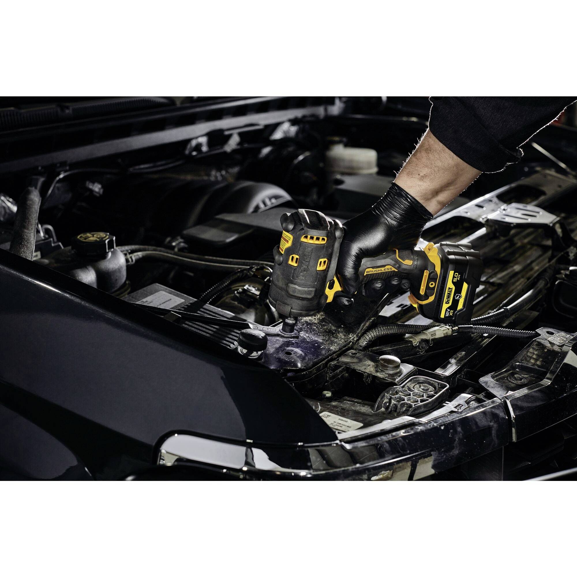 Eine Person verwendet einen Akkubohrer, um an einem offenen Motorraum eines Autos zu arbeiten. Die Hand trägt einen Handschuh.