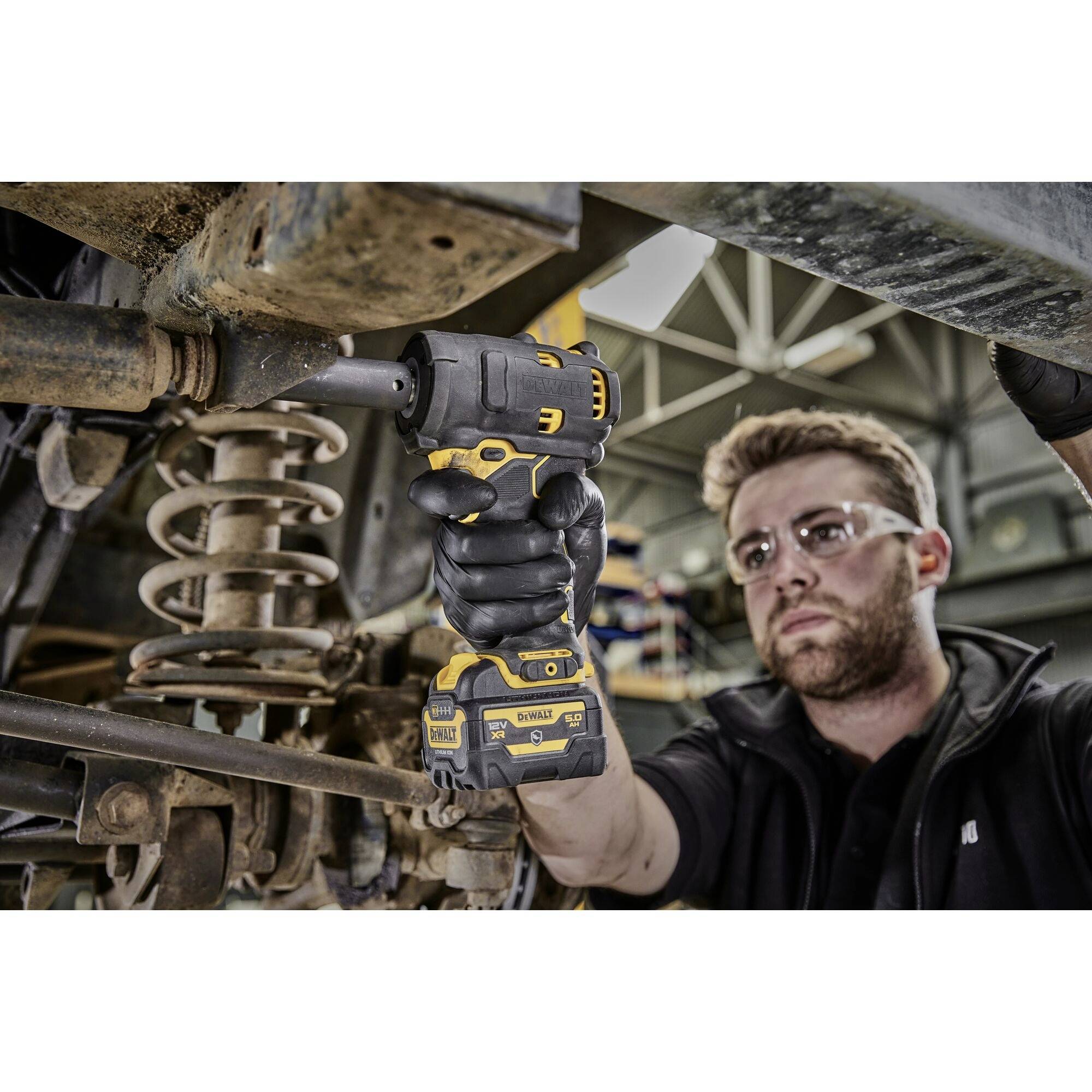 Eine Person mit Schutzbrille benutzt einen Akkuschrauber, um an einem Fahrzeugfahrwerk zu arbeiten. Im Hintergrund eine Werkstattumgebung.