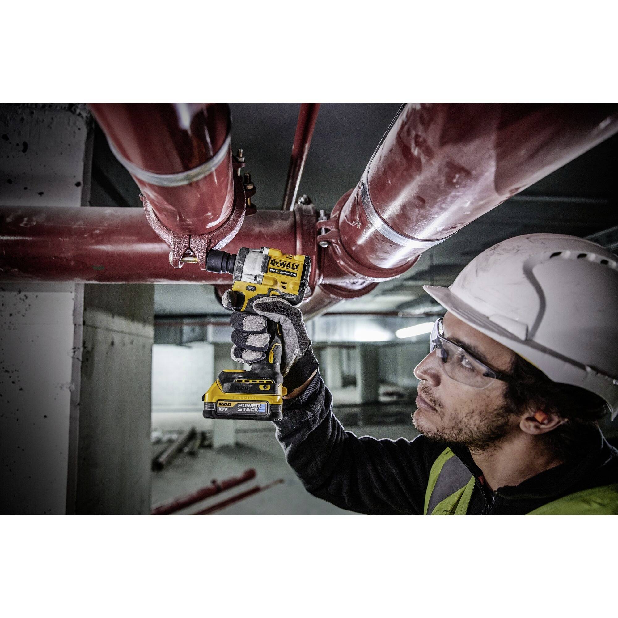 Ein Arbeiter mit Schutzhelm und Brille benutzt einen Akkuschrauber, um rote Rohre in einem Gebäude zu befestigen.