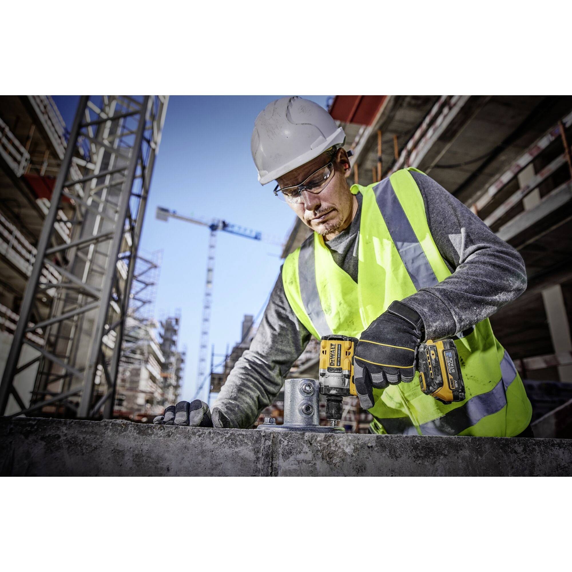 Ein Bauarbeiter in Schutzkleidung benutzt einen Bohrer auf einer Baustelle. Im Hintergrund sind Baukräne und teilweise fertiggestellte Gebäude zu sehen.