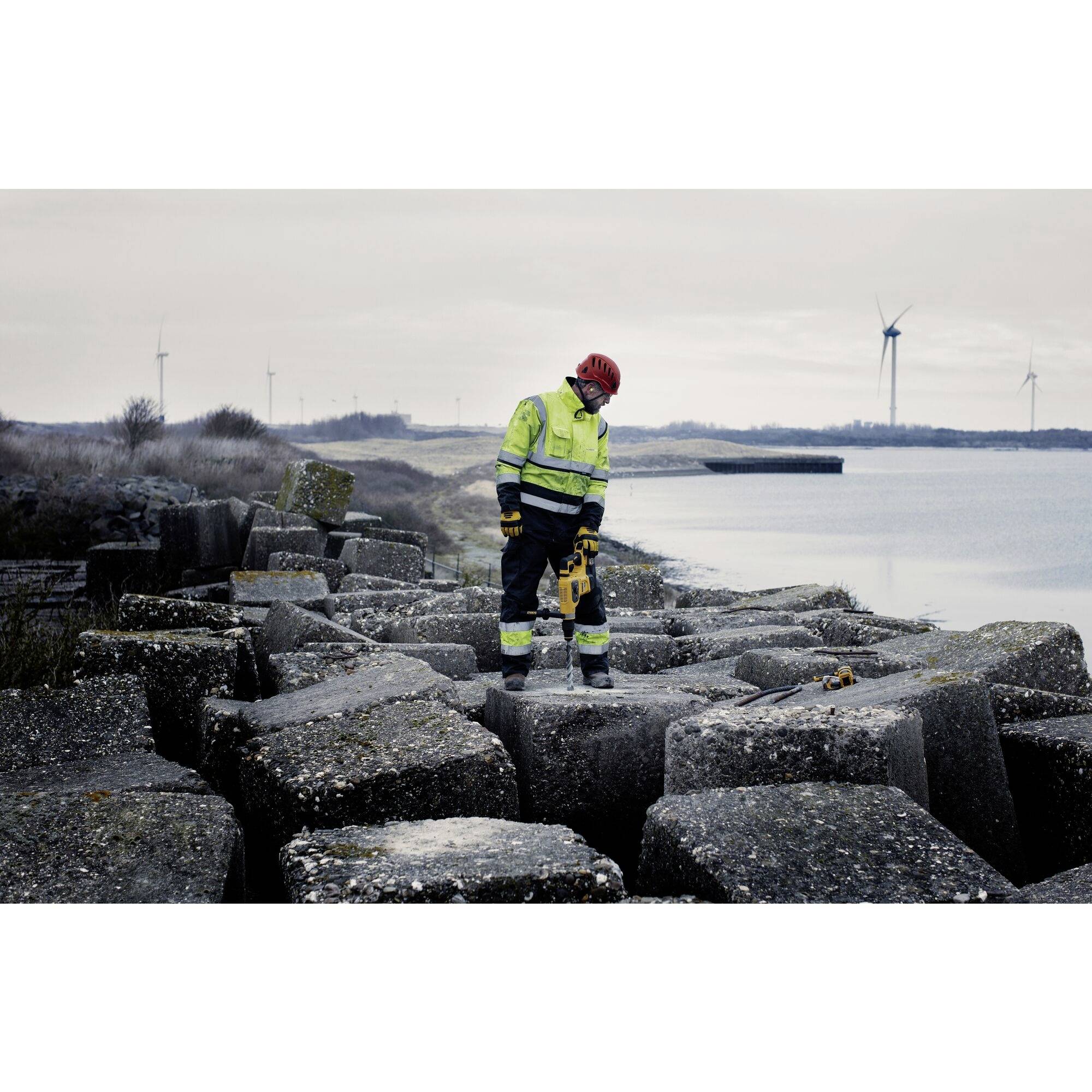Person im Schutzanzug mit Helden steht auf großen Betonblöcken nahe einem Gewässer, im Hintergrund Windkraftanlagen und bewölkter Himmel.