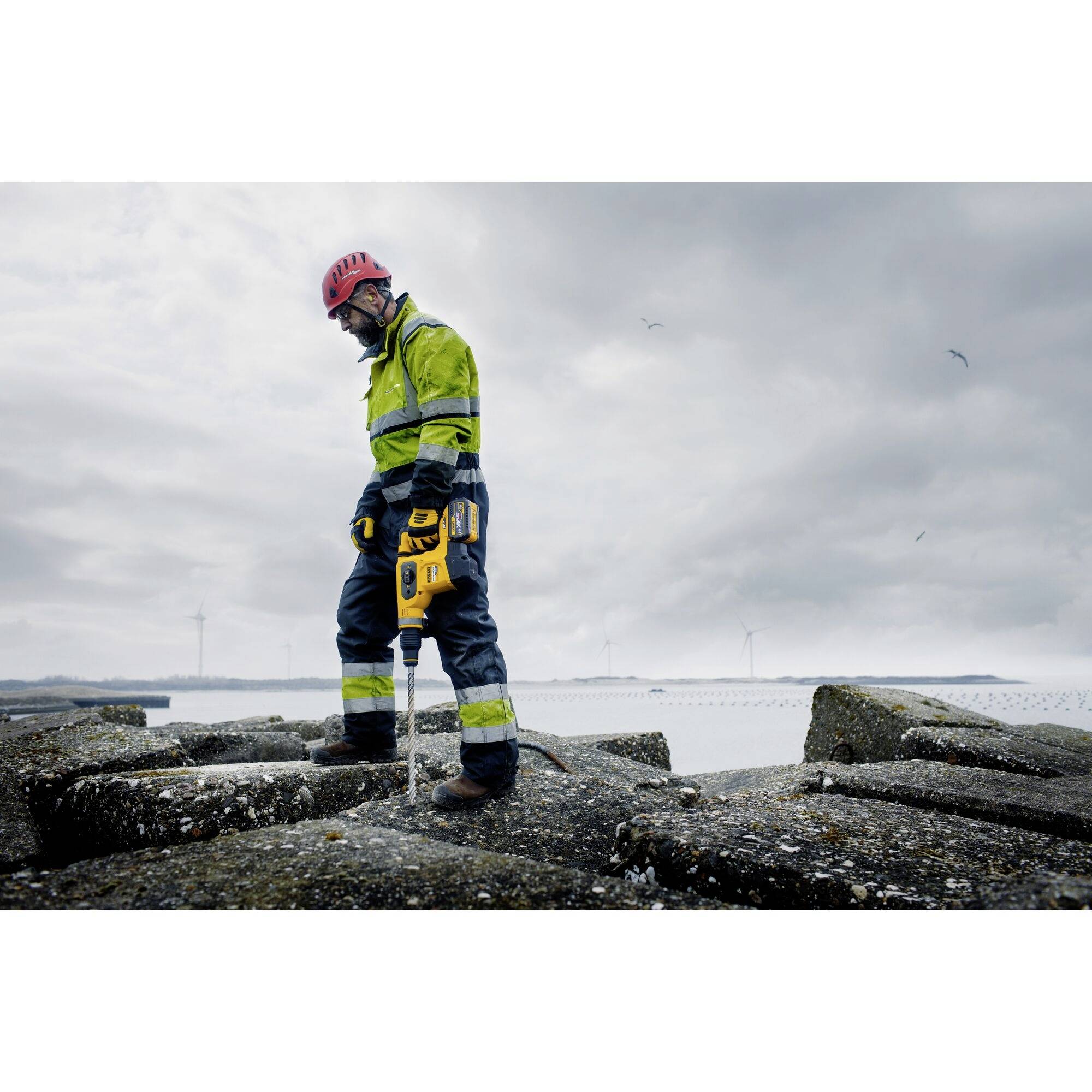 Ein Arbeiter in Sicherheitskleidung mit Helm steht auf felsigem Untergrund und bohrt mit einem gelben Gerät. Im Hintergrund Windkraftanlagen.