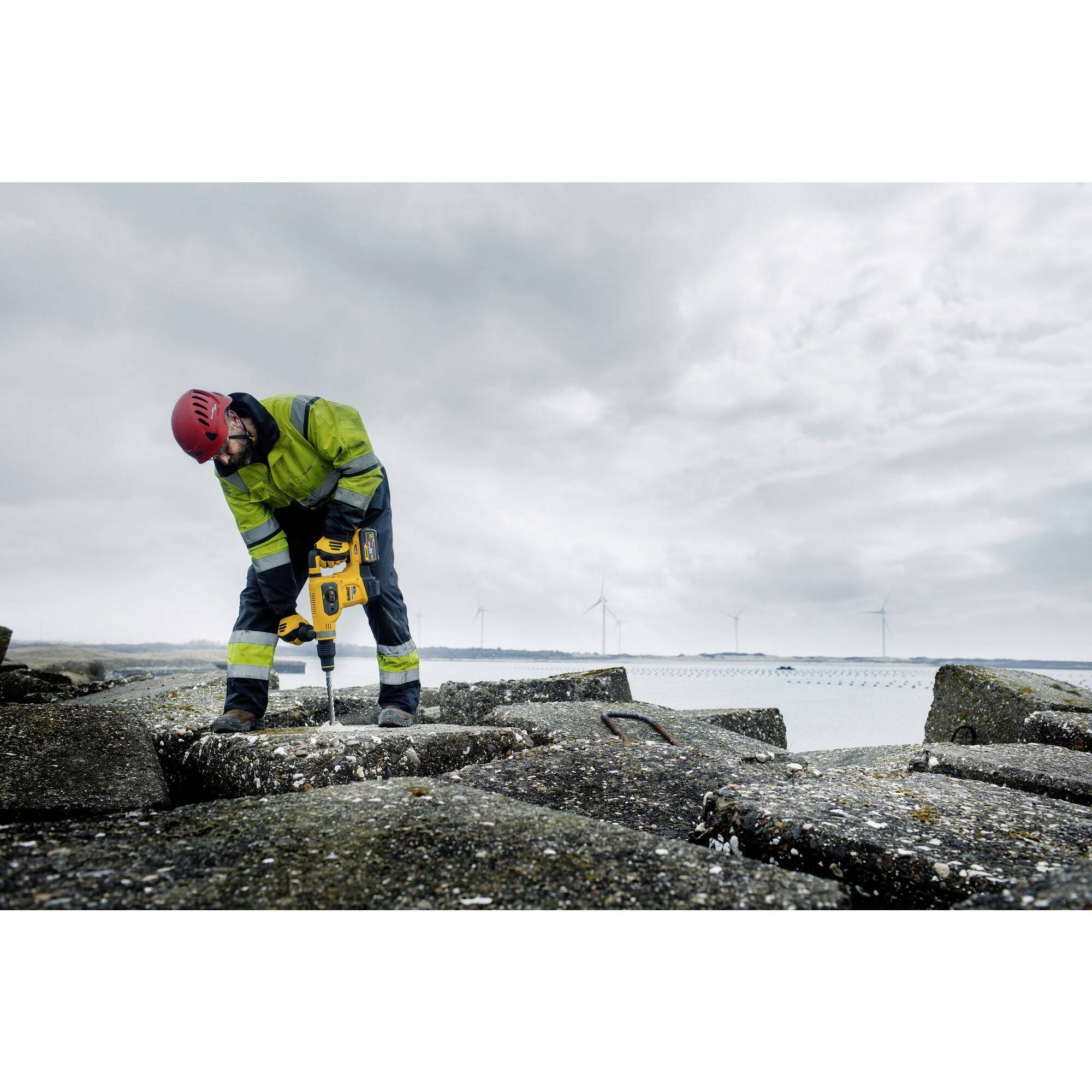 Ein Arbeiter mit rotem Helm und Sicherheitskleidung bohrt mit einer Maschine in eine Betonfläche. Im Hintergrund sind Windkraftanlagen sichtbar.