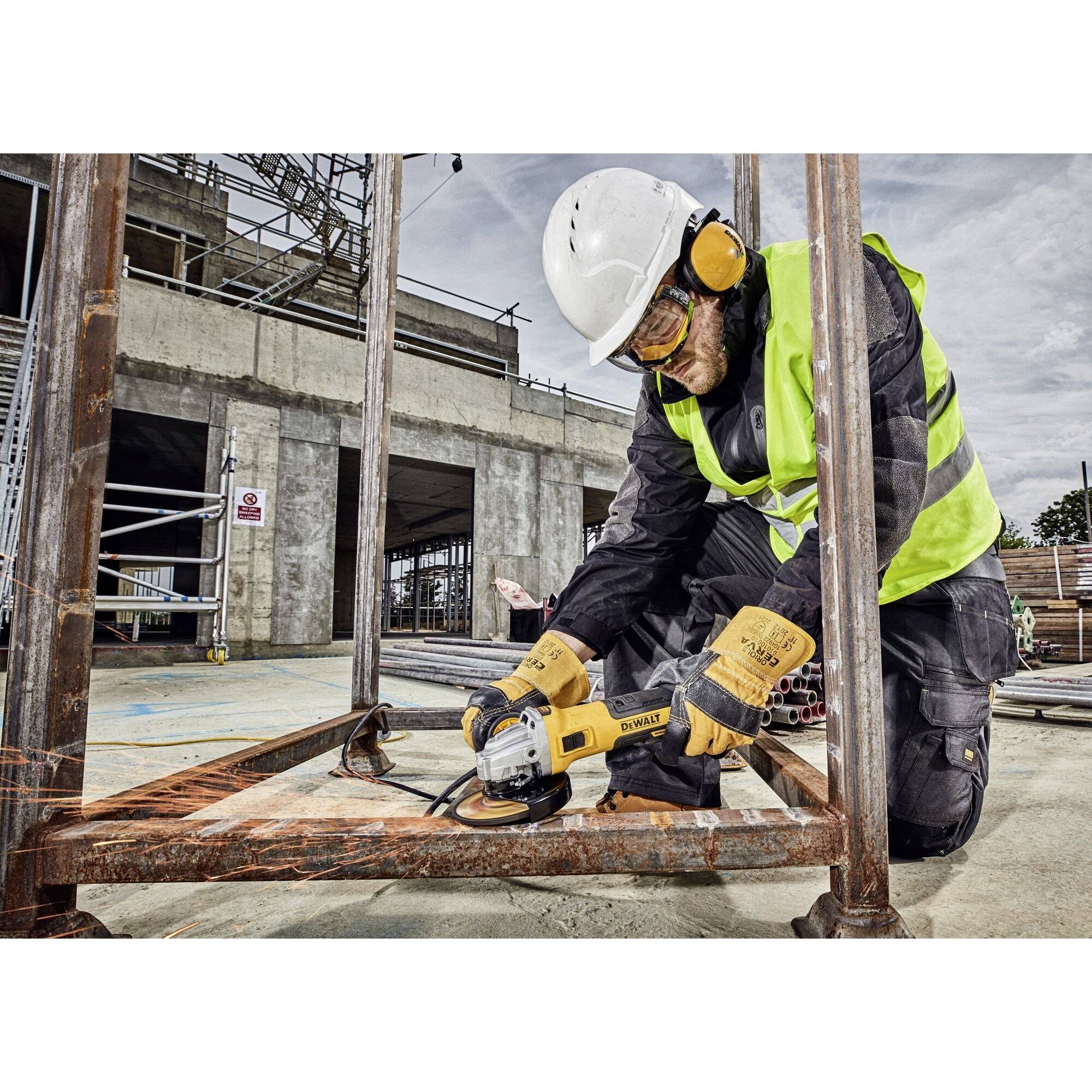 Arbeiter mit Schutzhelm und -kleidung schleift Metall mit Winkelschleifer auf Baustelle. Umgebung mit Baugerüst und Rohbau.