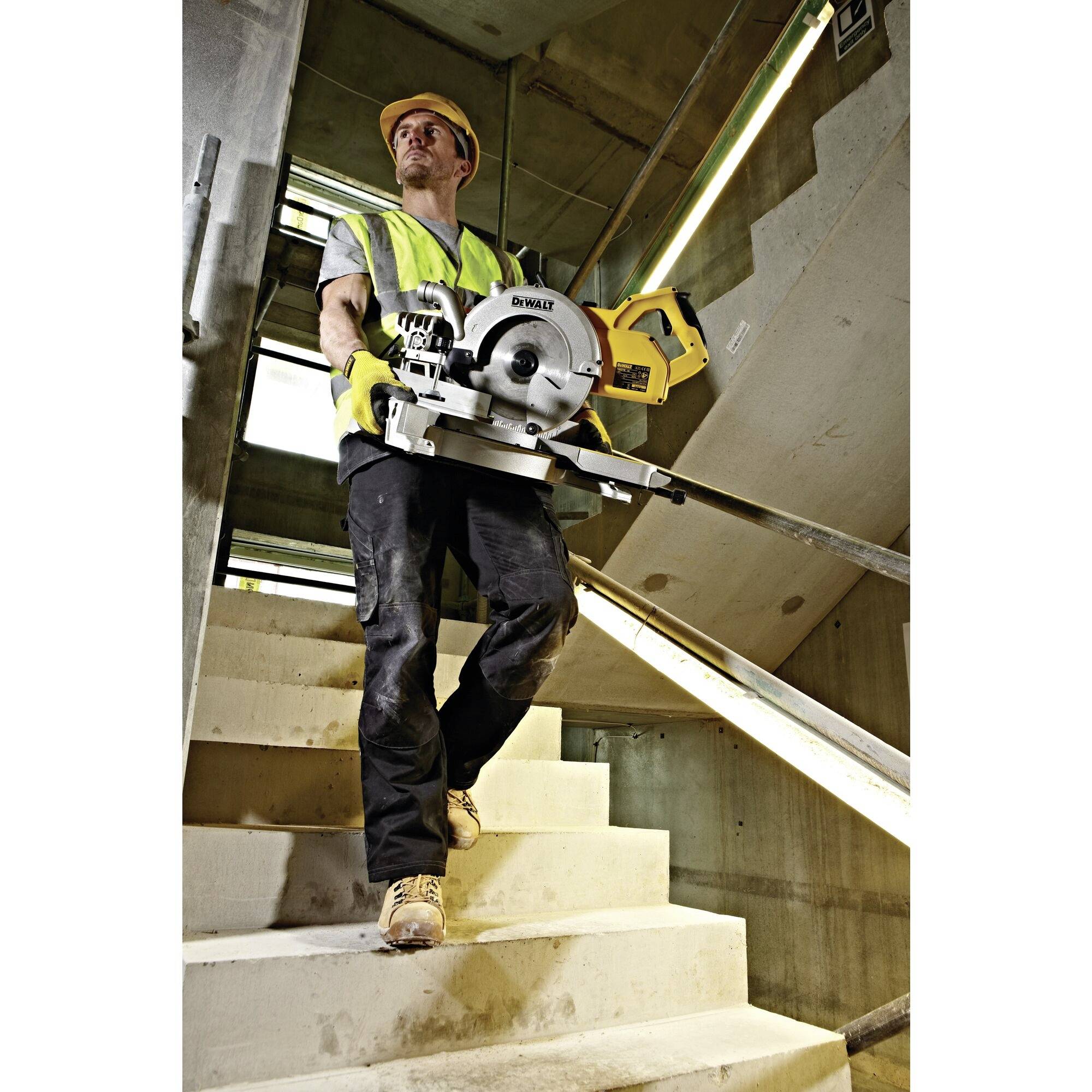 Arbeiter in Schutzkleidung trägt eine Kreissäge auf einer Baustellentreppe.
