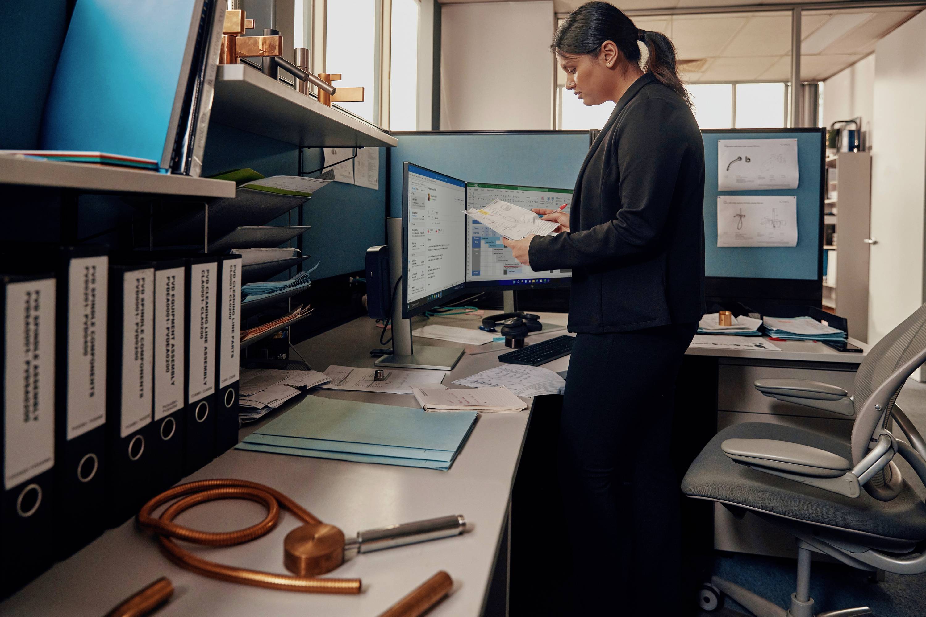 Eine Person im Büro überprüft Dokumente und betrachtet einen Computerbildschirm. Der Schreibtisch ist mit Ordnern und Papierstapeln bedeckt.