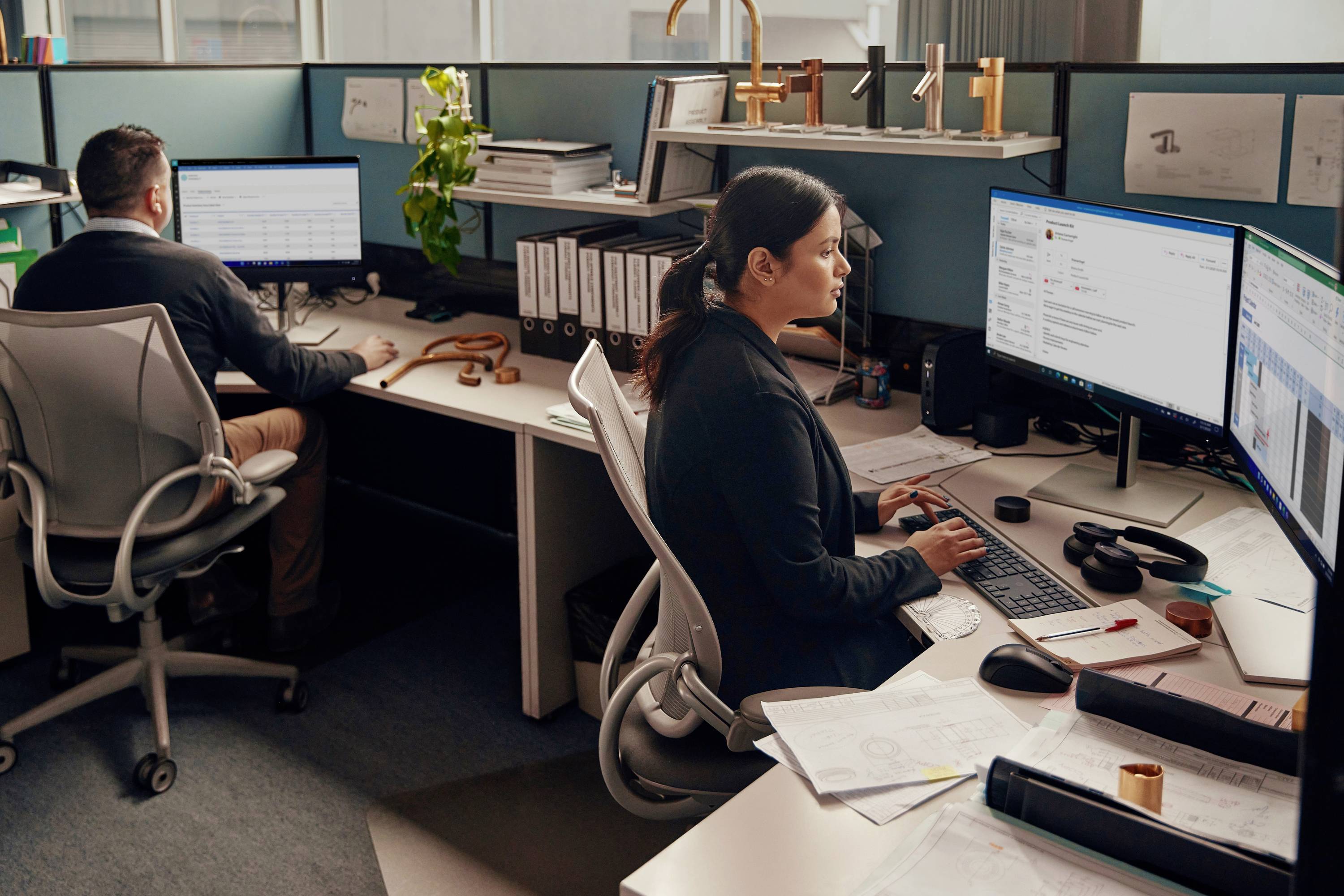 Zwei Personen arbeiten in einem Büro an Computern. Beide sitzen an Schreibtischen mit Monitoren, Ordnern und Papieren.