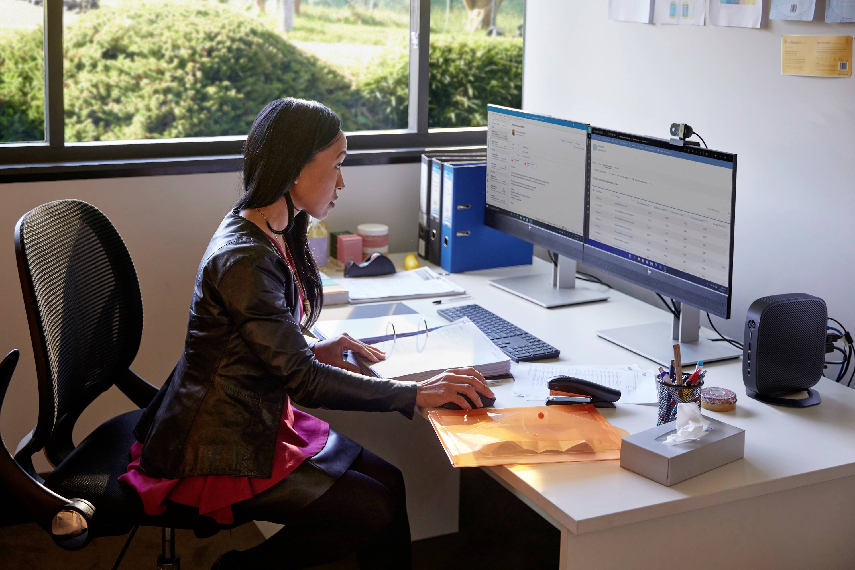 Eine Frau sitzt an einem Schreibtisch und arbeitet am Computer. Sie trägt eine schwarze Jacke und ein pinkfarbenes Kleid. Im Hintergrund sind Pflanzen und Ordner zu sehen.