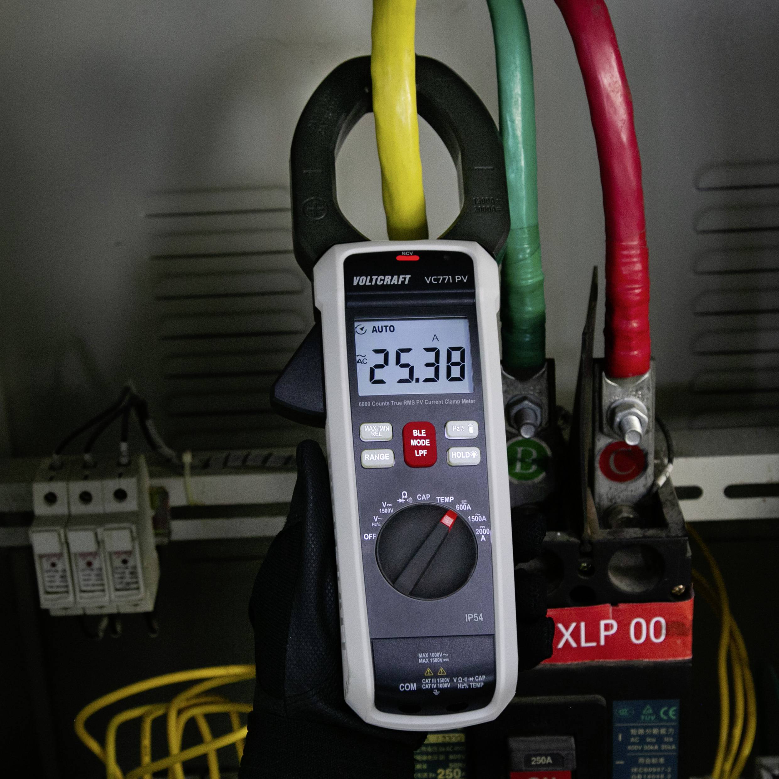 Ein Multimeter misst die Spannung und zeigt '25,38' auf einer elektrischen Schaltanlage mit gelben, grünen und roten Kabeln an.