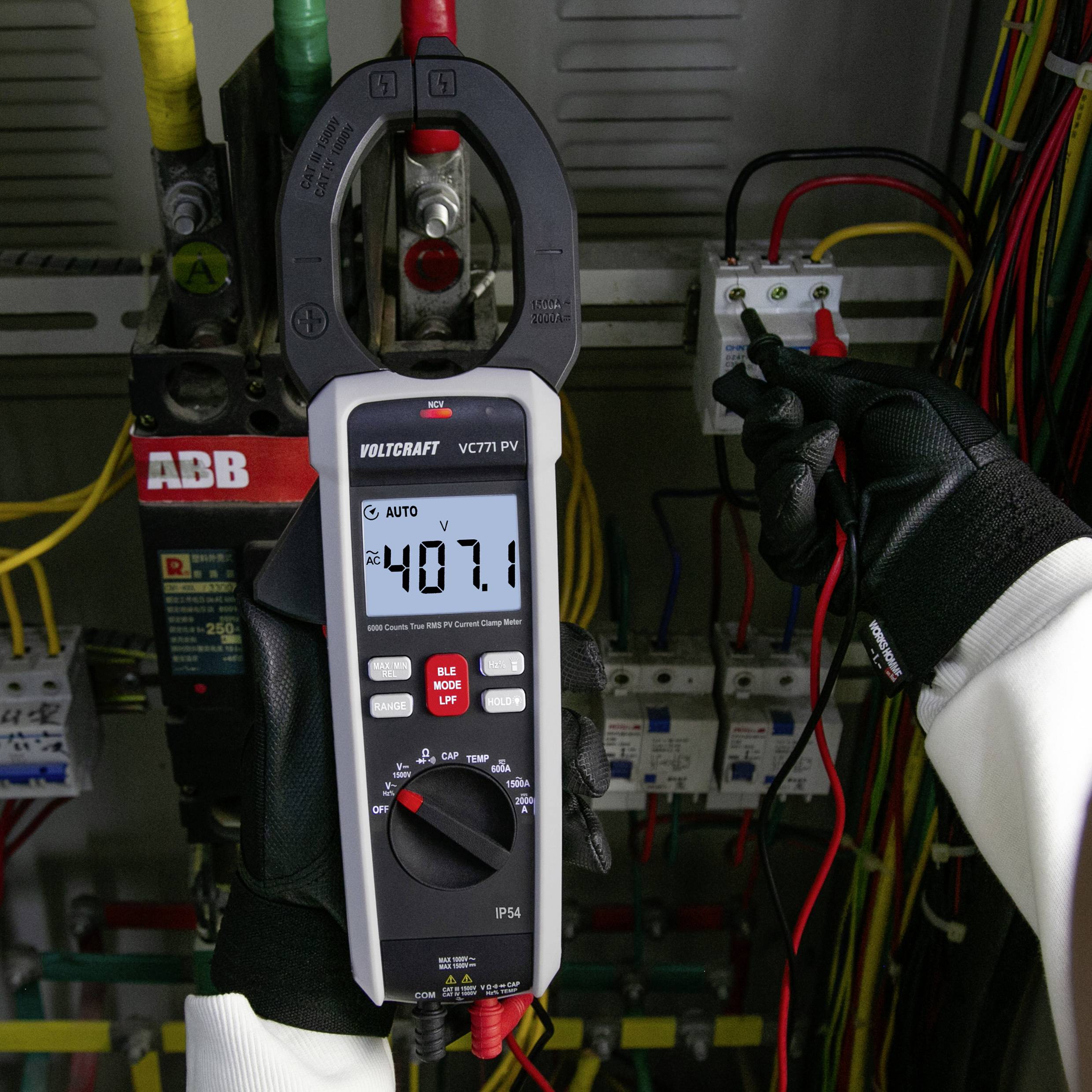 Eine Person mit Handschuhen misst mit einem Multimeter die Spannung und zeigt '407,1' auf dem Display vor einem Kontrollpanel mit Kabeln.