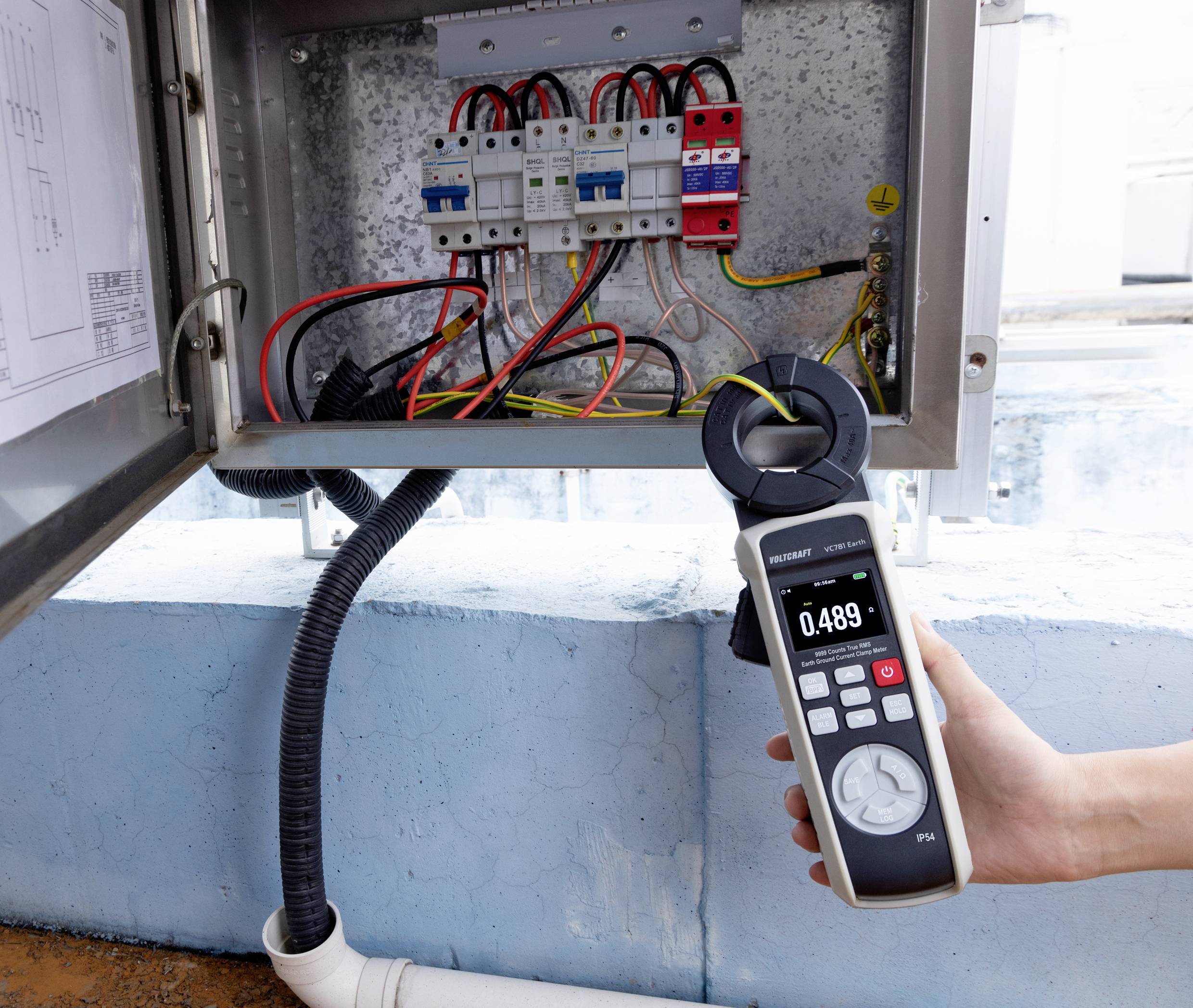 Hand, die ein digitales Multimeter mit der Anzeige 0,489 in der Nähe einer Elektroverteilerdose mit sichtbaren Kabeln und Sicherungen hält, blauer Wandhintergrund.