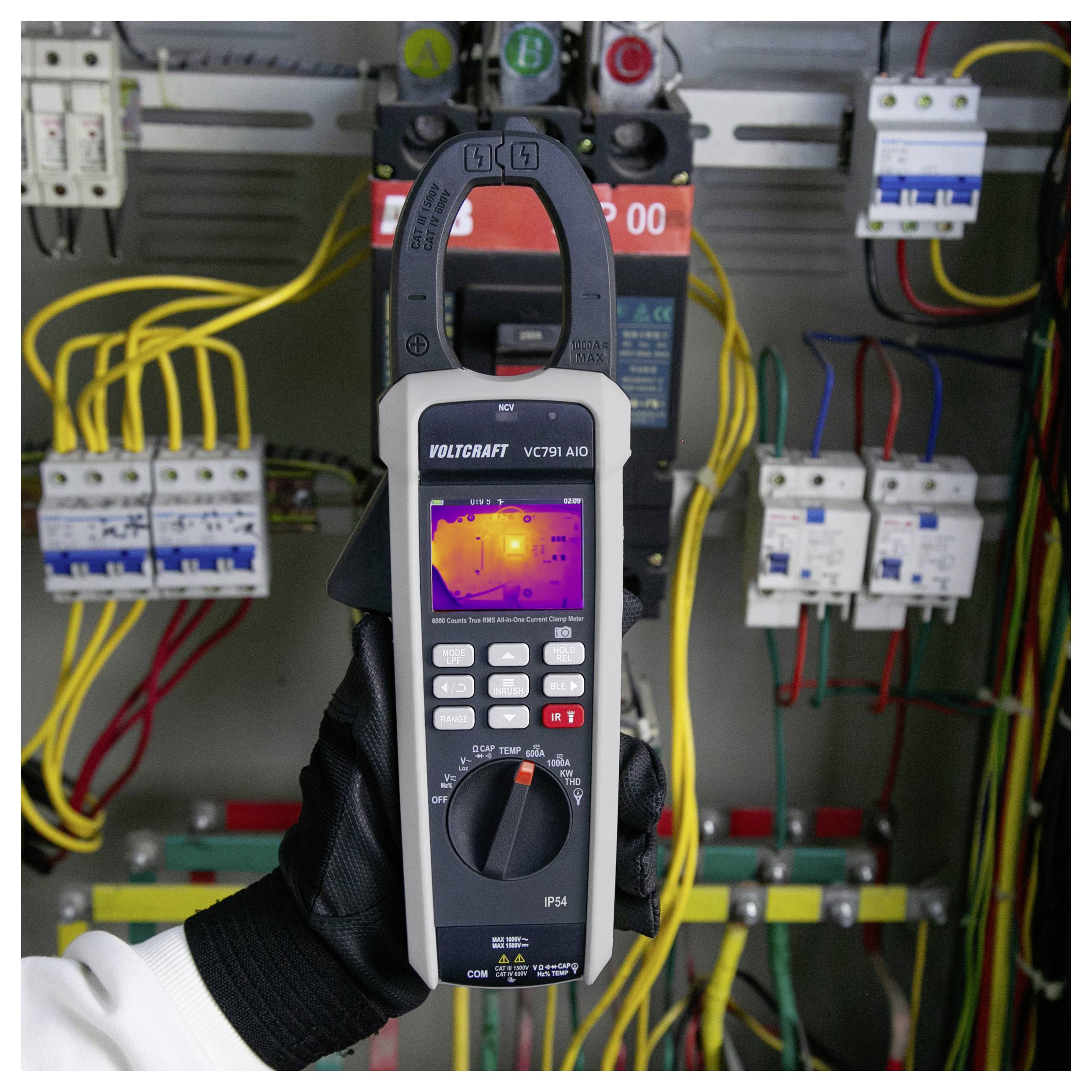 Eine behandschuhte Hand hält ein VOLTCRAFT-Zangenmessgerät mit Wärmebild auf einem Bildschirm im Inneren eines elektrischen Schaltschranks mit Drähten und Schutzschaltern.