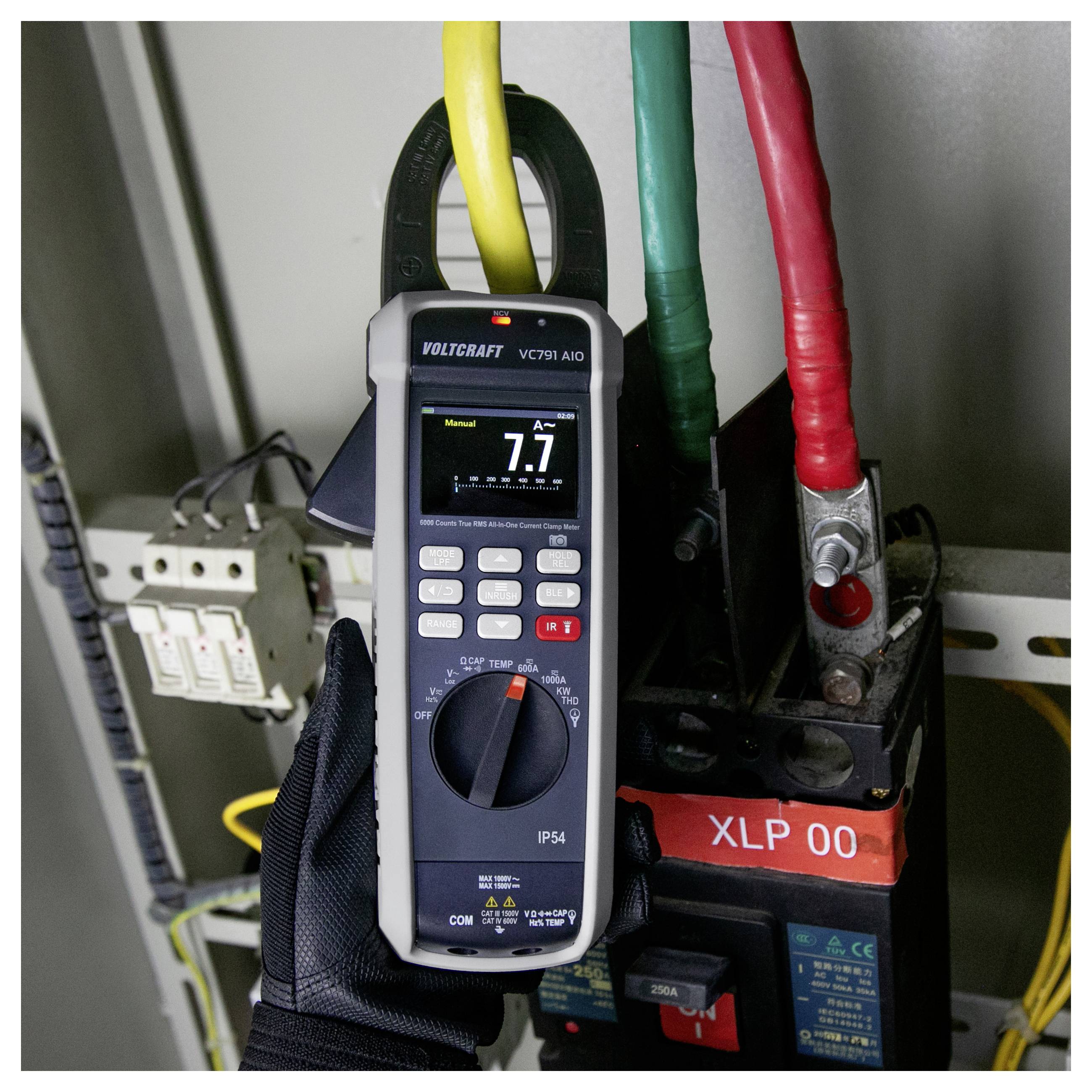 Zangenprüfer zeigt 7,7 A an, angebracht um ein gelbes Kabel in einem Elektroverteilerschrank mit roten und grünen Leitern und Schaltgeräten.