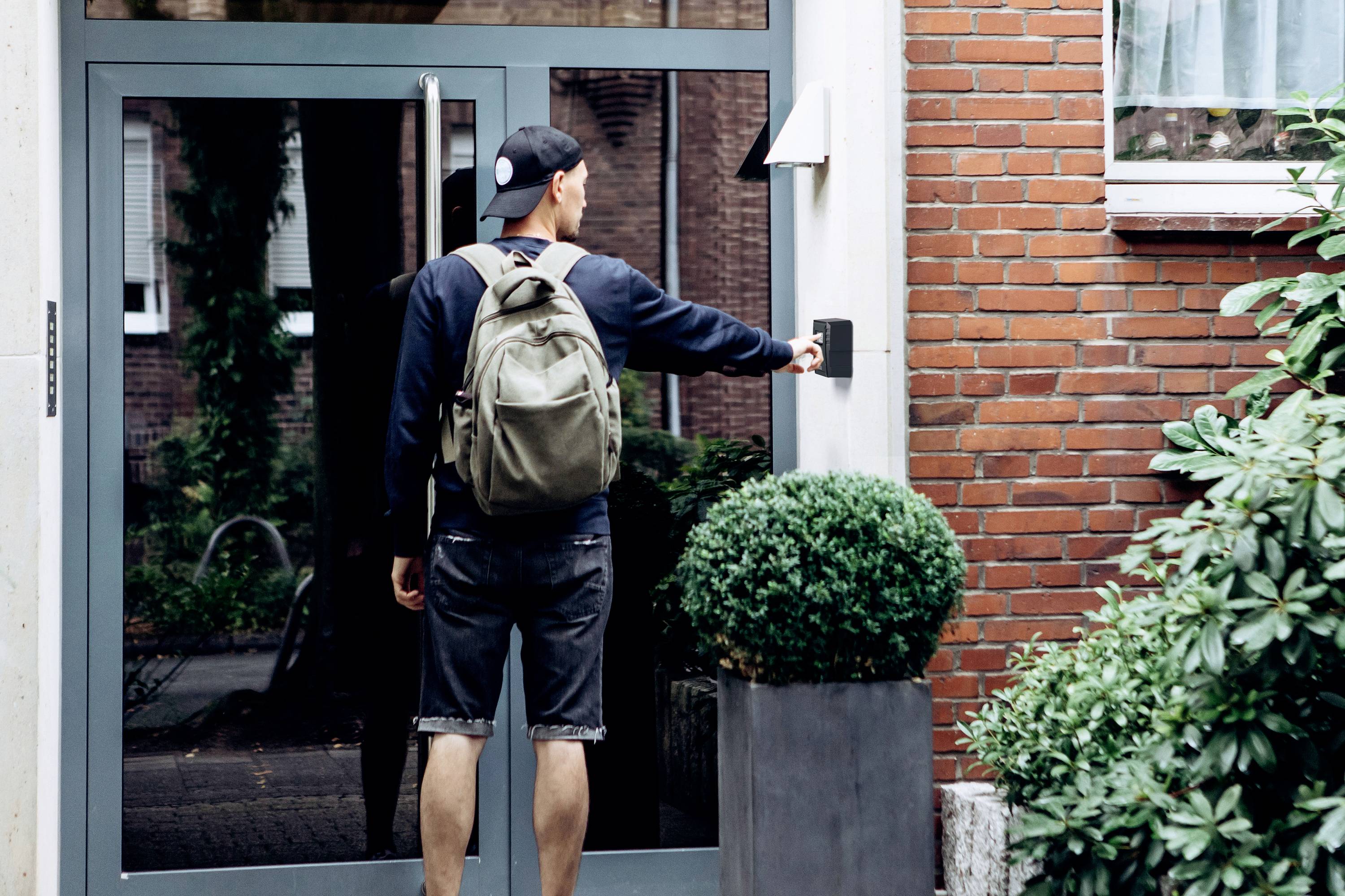 Person mit Rucksack klingelt an einer Haustür neben einer Ziegelmauer. Im Vordergrund sind Topfpflanzen sichtbar.