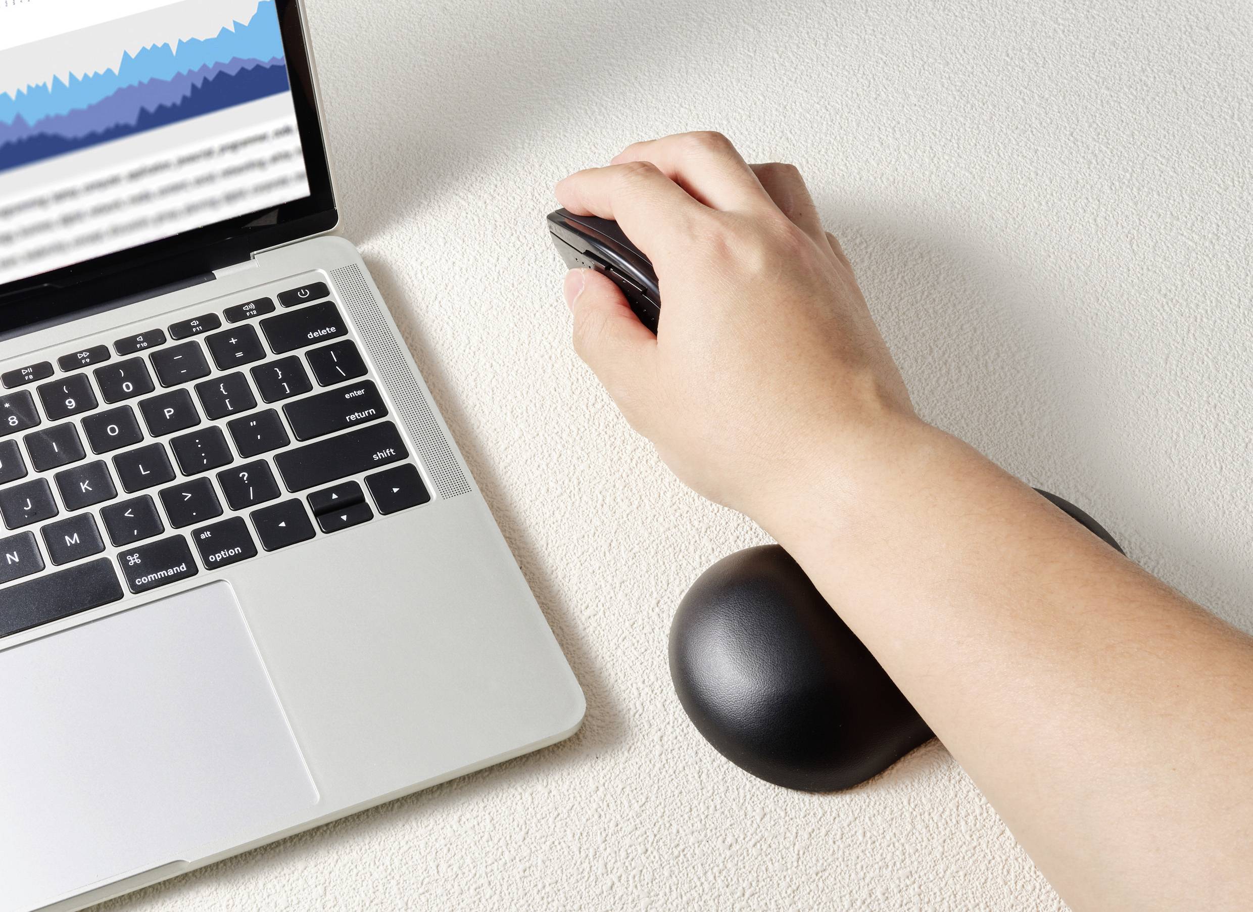 Eine Person arbeitet am Laptop mit einer ergonomischen Handgelenkauflage und hält eine Maus. Der Laptopbildschirm zeigt Grafiken.