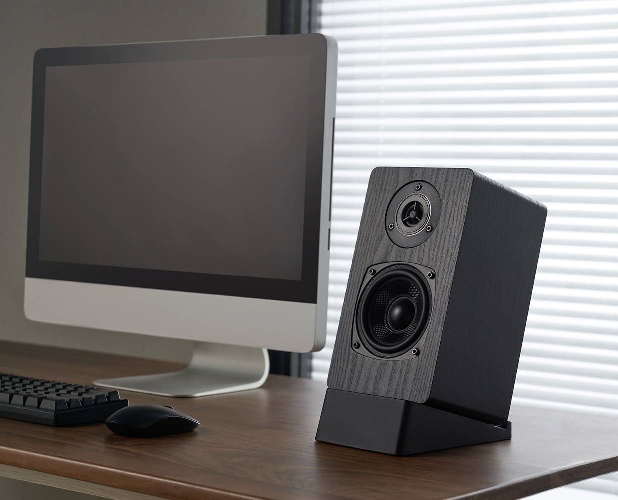 Ein Computer mit Monitor und Tastatur auf einem Holzschreibtisch. Rechts steht ein schwarzer Lautsprecher. Im Hintergrund Jalousien.