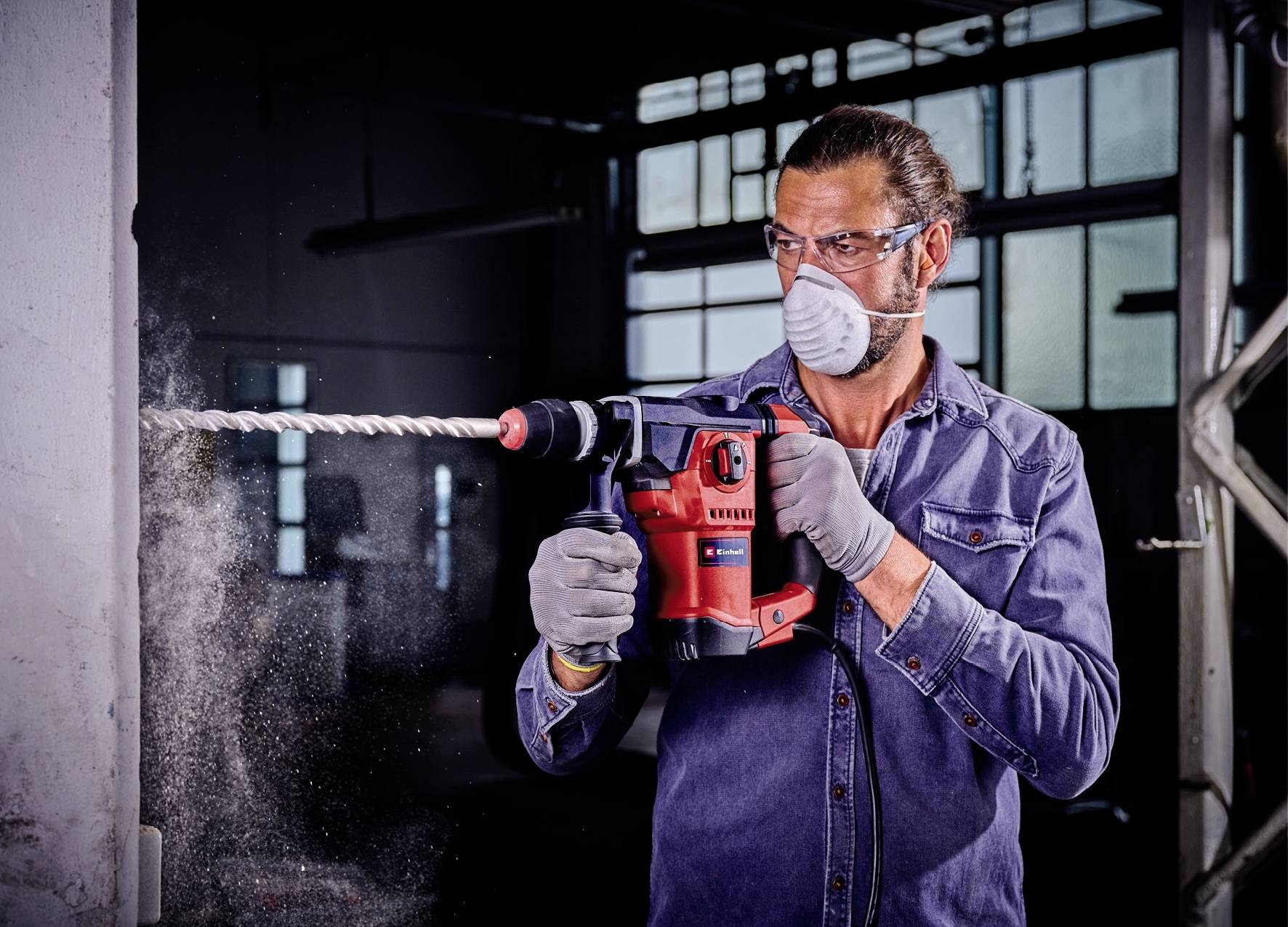 Mann in blauer Jeansjacke und Schutzmaske bohrt mit einer roten Bohrmaschine in eine Betonwand, Staub fliegt heraus.