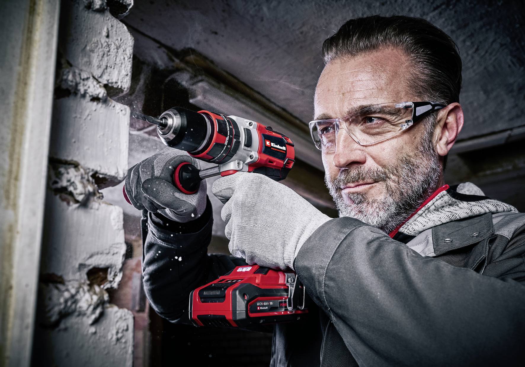 Ein Mann trägt Schutzbrille und Handschuhe, während er eine rote und schwarze Bohrmaschine zur Arbeit an einer weißen Ziegelwand verwendet.