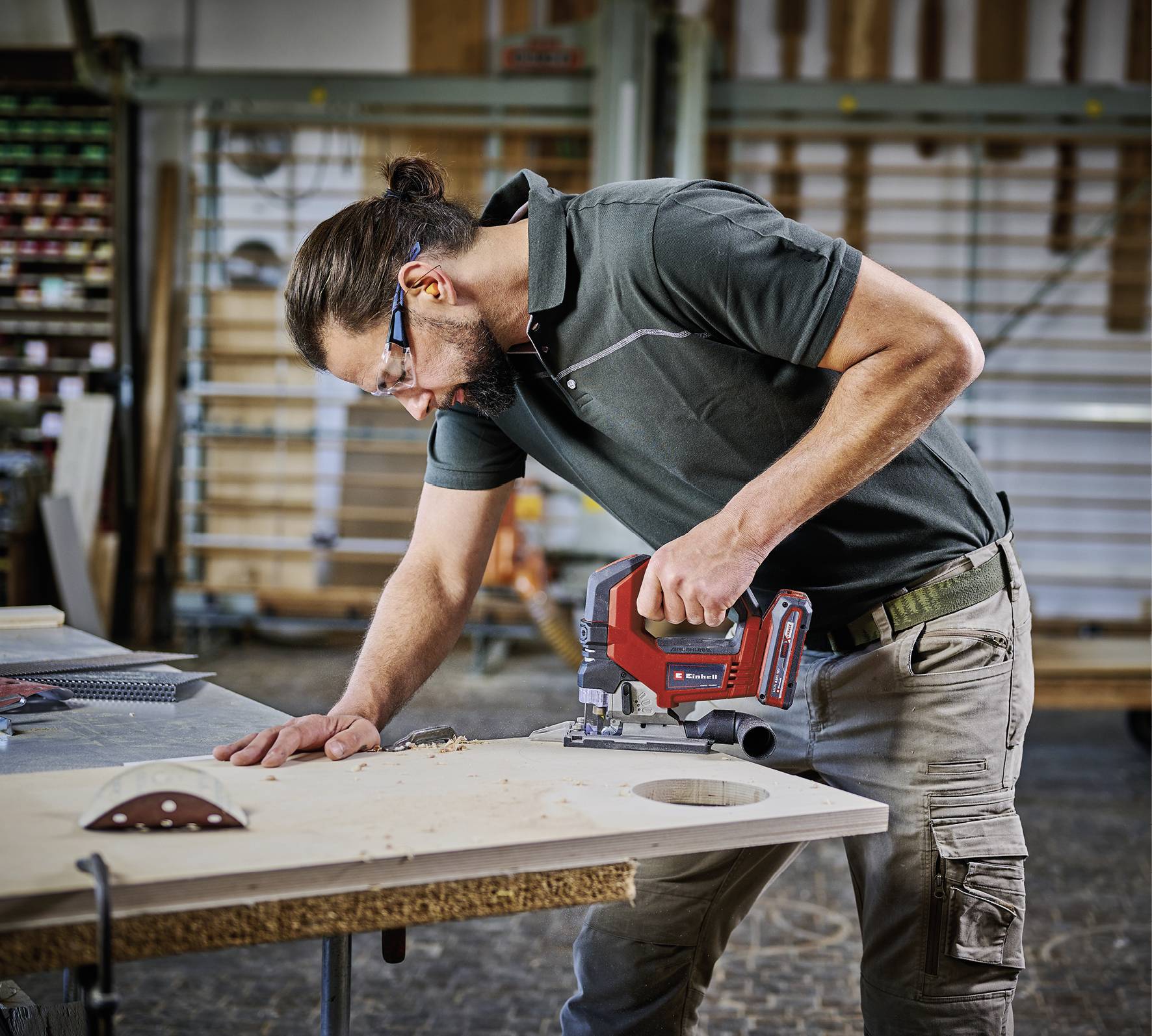 Eine Person mit Sicherheitsbrille und Poloshirt verwendet eine elektrische Säge, um in einer Werkstattumgebung ein rundes Loch in ein Holzbrett zu schneiden.