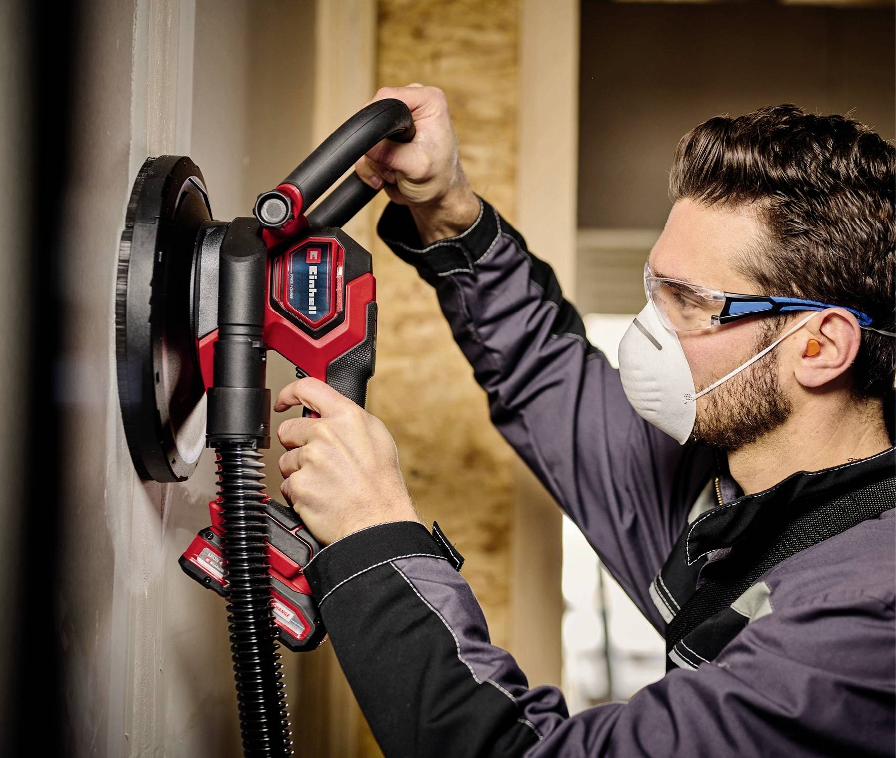Ein Mann trägt eine Schutzbrille und Maske und schleift mit einer elektrischen Schleifmaschine eine Wand.