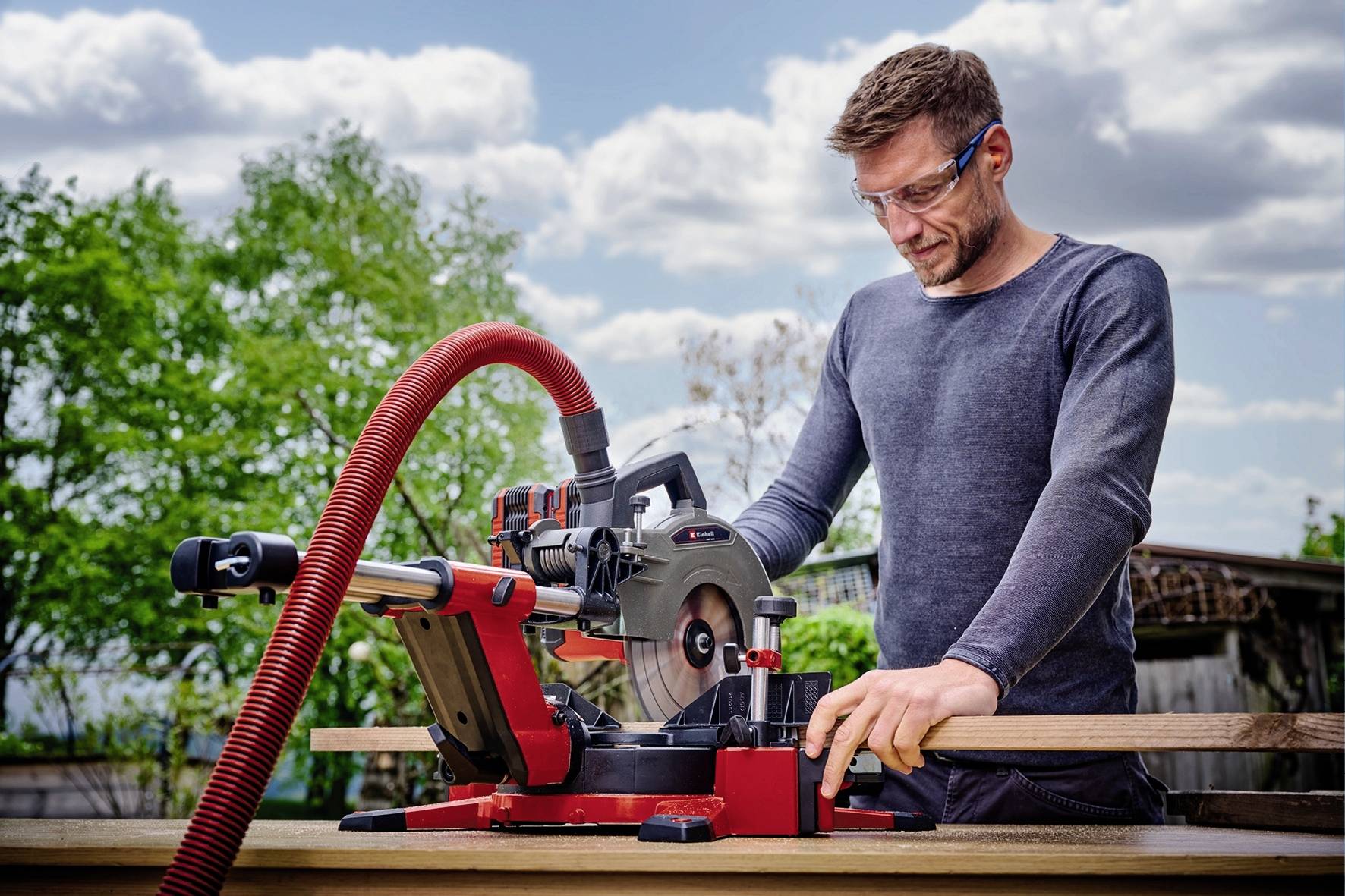Ein Mann mit Schutzbrille arbeitet im Freien an einer rot-schwarzen Kappsäge, umgeben von grünen Bäumen und bewölktem Himmel.