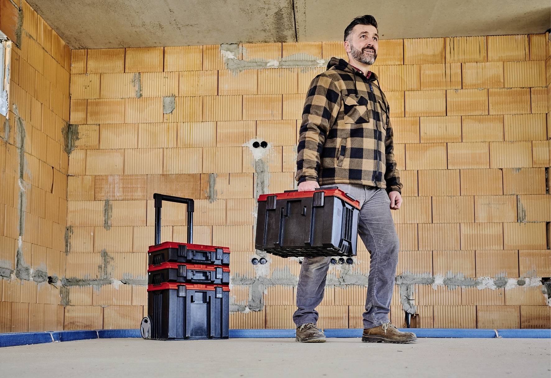 Ein Mann in Arbeitskleidung trägt einen Werkzeugkasten in einem unfertigen Raum. Neben ihm steht ein Rollwagen mit weiteren Kisten.