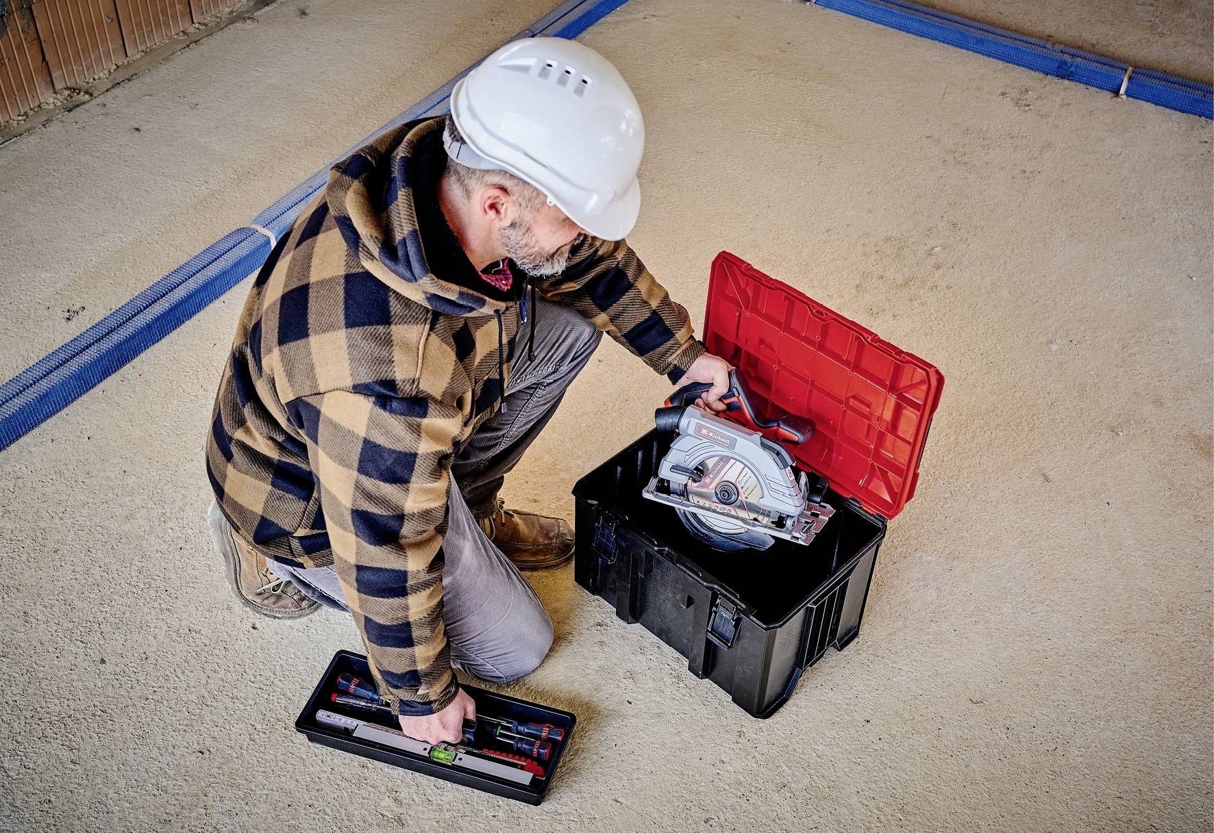 Eine Person trägt einen Schutzhelm und kniet auf dem Boden, öffnet eine Werkzeugkiste und holt eine Kreissäge heraus.