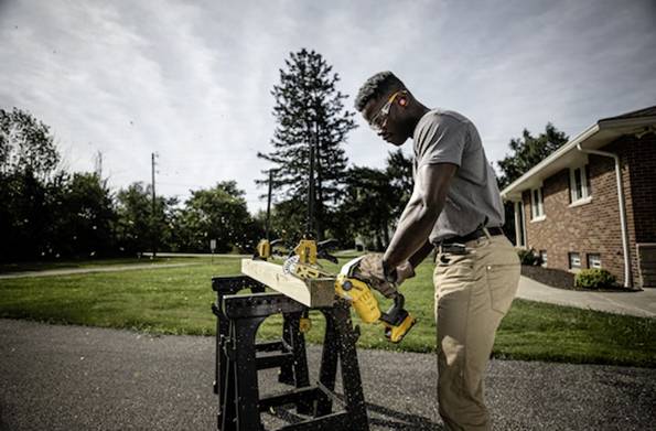 Eine Person, die draußen auf einem Sägebock Holz mit einer Kreissäge schneidet; trägt Schutzbrille, mit Bäumen und einem Ziegelhaus im Hintergrund.