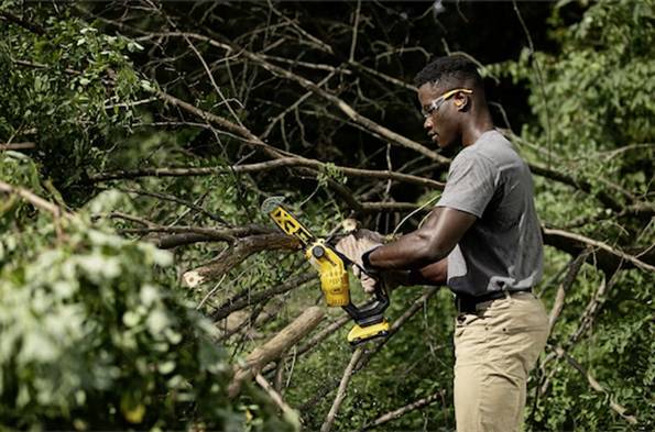 Eine Person mit Schutzbrille verwendet eine gelbe Kettensäge, um Äste in einem dichten, bewaldeten Gebiet zu schneiden.