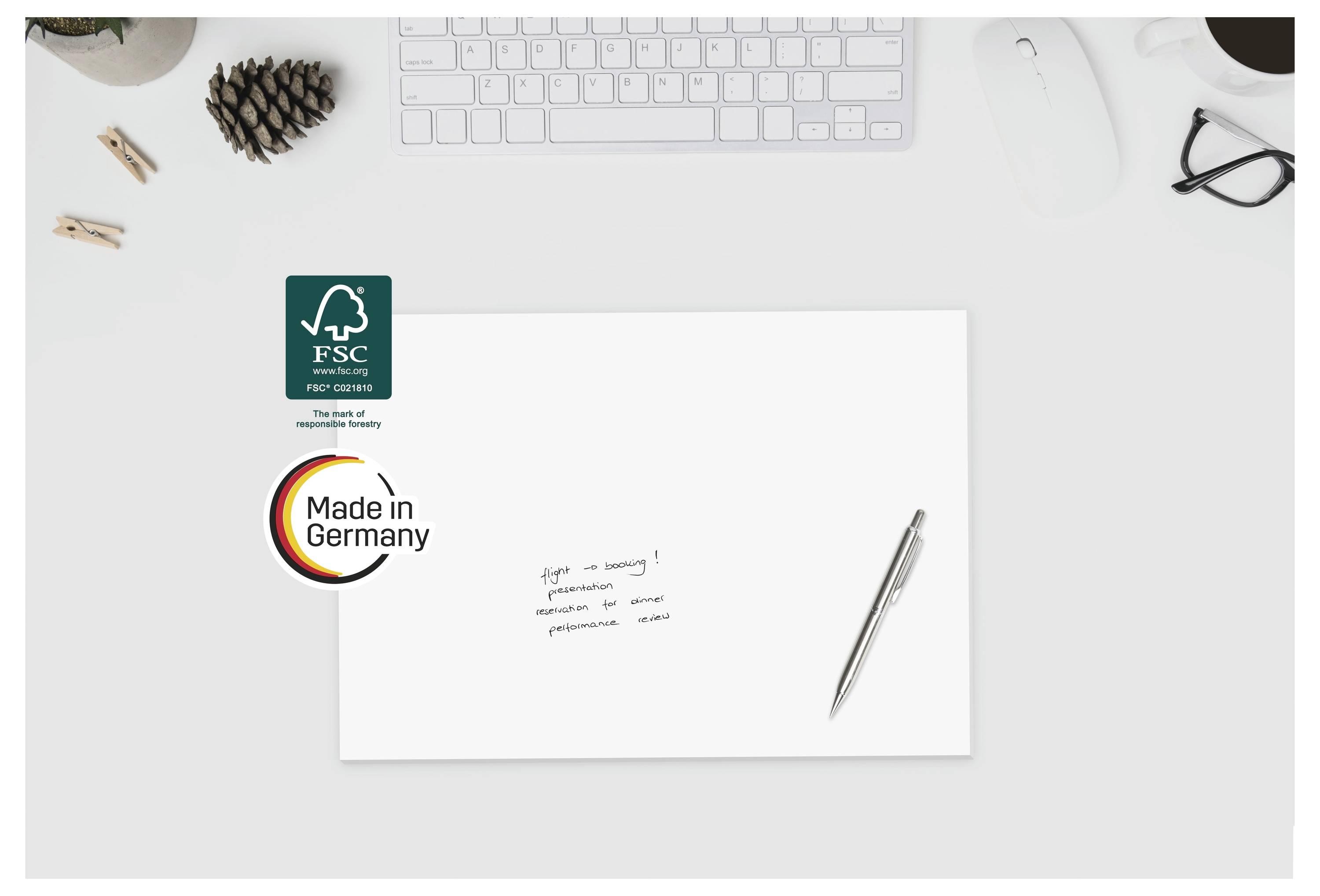 Schreibtischszene mit einer leeren weißen Karte oder einem Umschlag, die FSC- und „Made in Germany"-Logos zeigen, einem Stift daneben und einer Tastatur im Hintergrund.