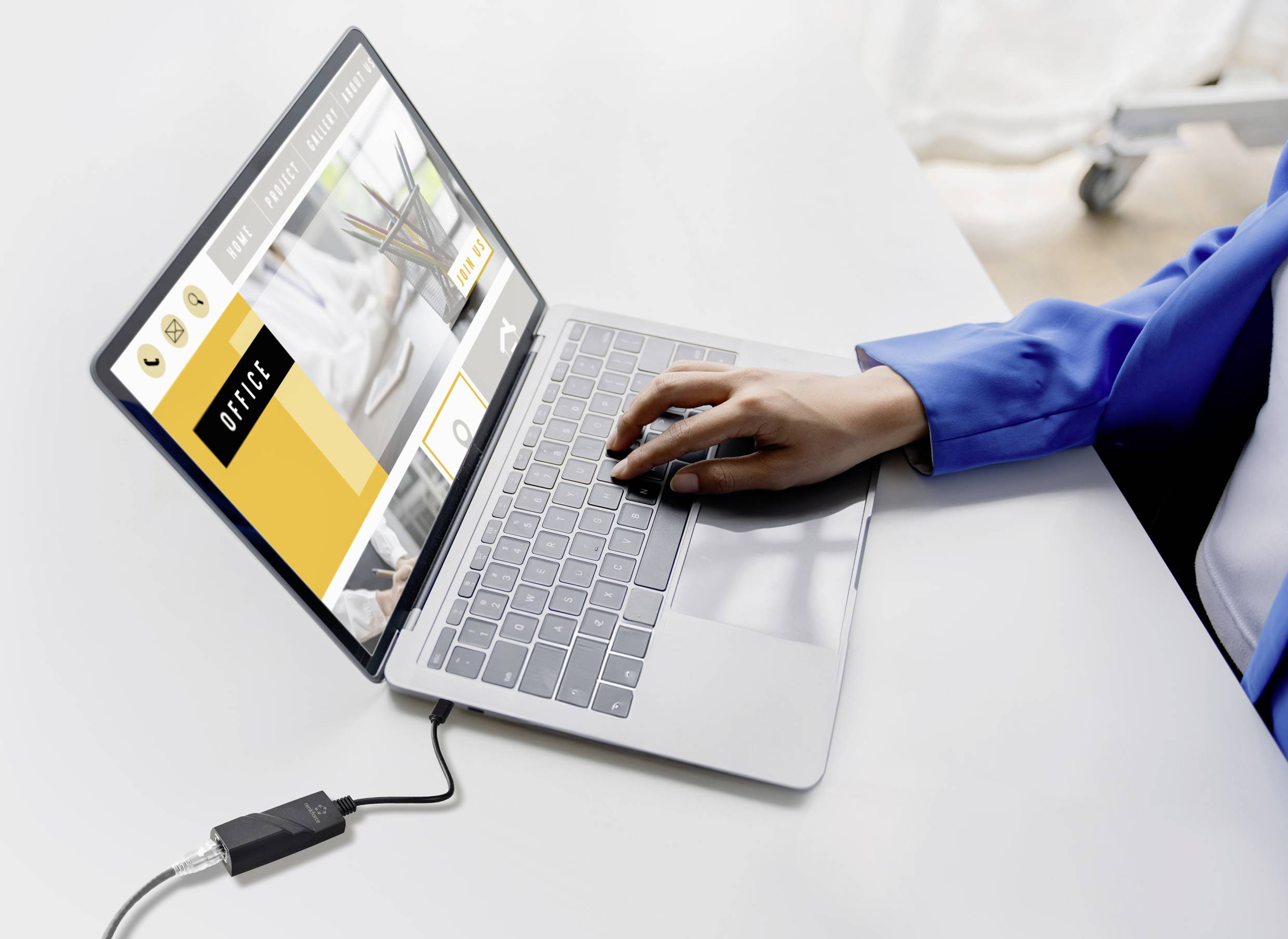 Eine Person in blauem Blazer tippt auf einem Laptop mit geöffneter Webseite. Der Laptop ist mit einem Adapter verbunden.