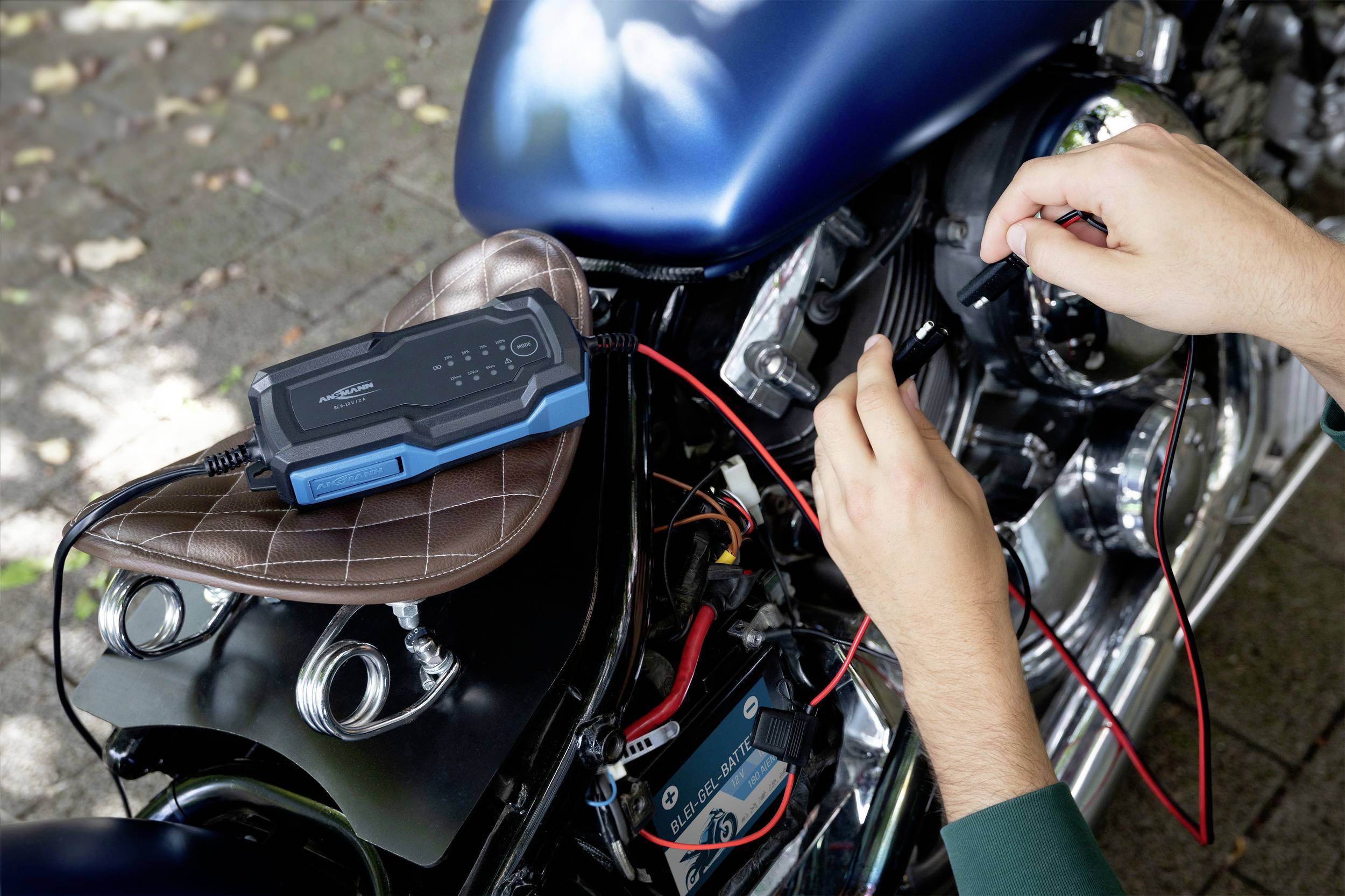 Ein Ladegerät ist mit einem Motorrad verbunden. Zwei Hände halten die Kabel an den Batterieklemmen. Der Sitz ist braun.