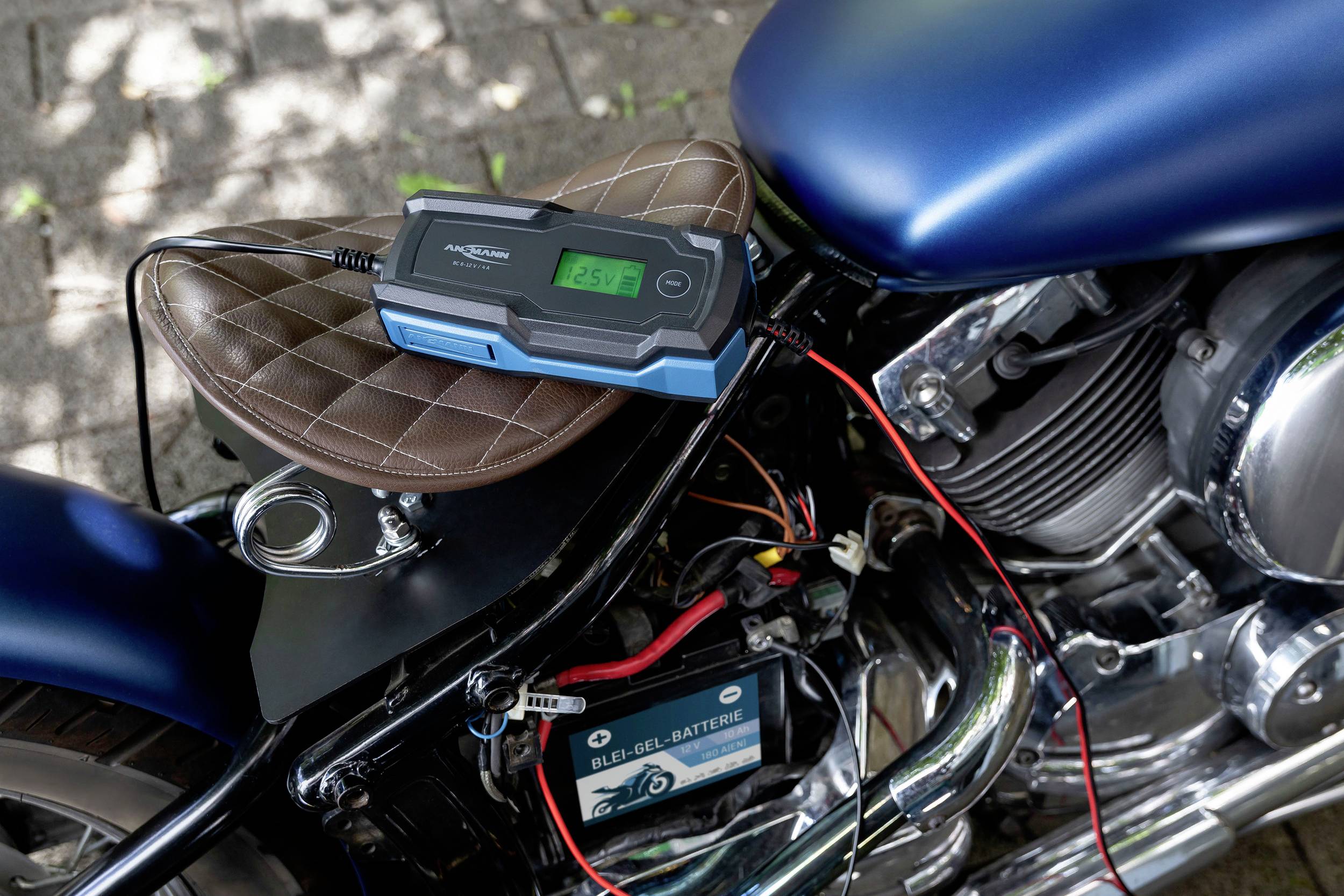 Ein Motorrad mit einer angeschlossenen Batterie und einem Ladegerät, das auf dem Sitz liegt. Das Display zeigt den Ladestatus an.